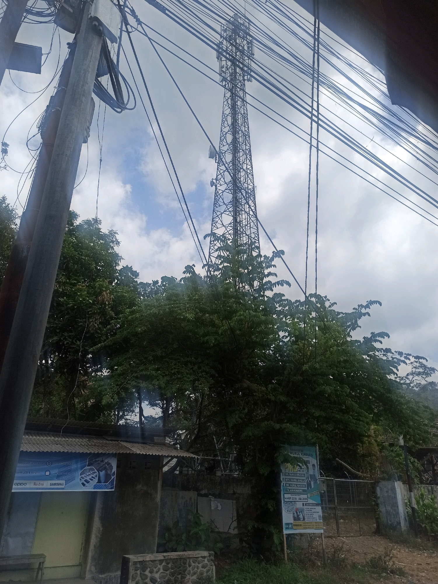 The image depicts an outdoor scene with a prominent cell tower in the background, a utility pole with numerous wires in the foreground, lush green trees, and a low-rise building with signage. The setting appears to be a somewhat rural or semi-urban area in Tlogosari, Indonesia, given the presence of overhead electrical and communication lines common in such locations. The sky is overcast with some patches of blue visible.
The cell tower is a tall, lattice-style structure with antennae at its apex. The utility pole is thick and grey, supporting a dense network of black wires that crisscross the frame. To the left, a simple, weathered building has a corrugated metal roof and a banner advertising mobile phone brands such as Oppo, Redmi, and Samsung, along with a general statement about selling phones and batteries. To the right of the building, there is a green fence and a white post with a sign. In the middle ground, dense foliage from trees obscures parts of the scene. The ground is visible in the foreground and appears to be a dirt or gravel path. There are no people or animals visible in the image. The overall impression is one of everyday infrastructure in a developing or transitional area.