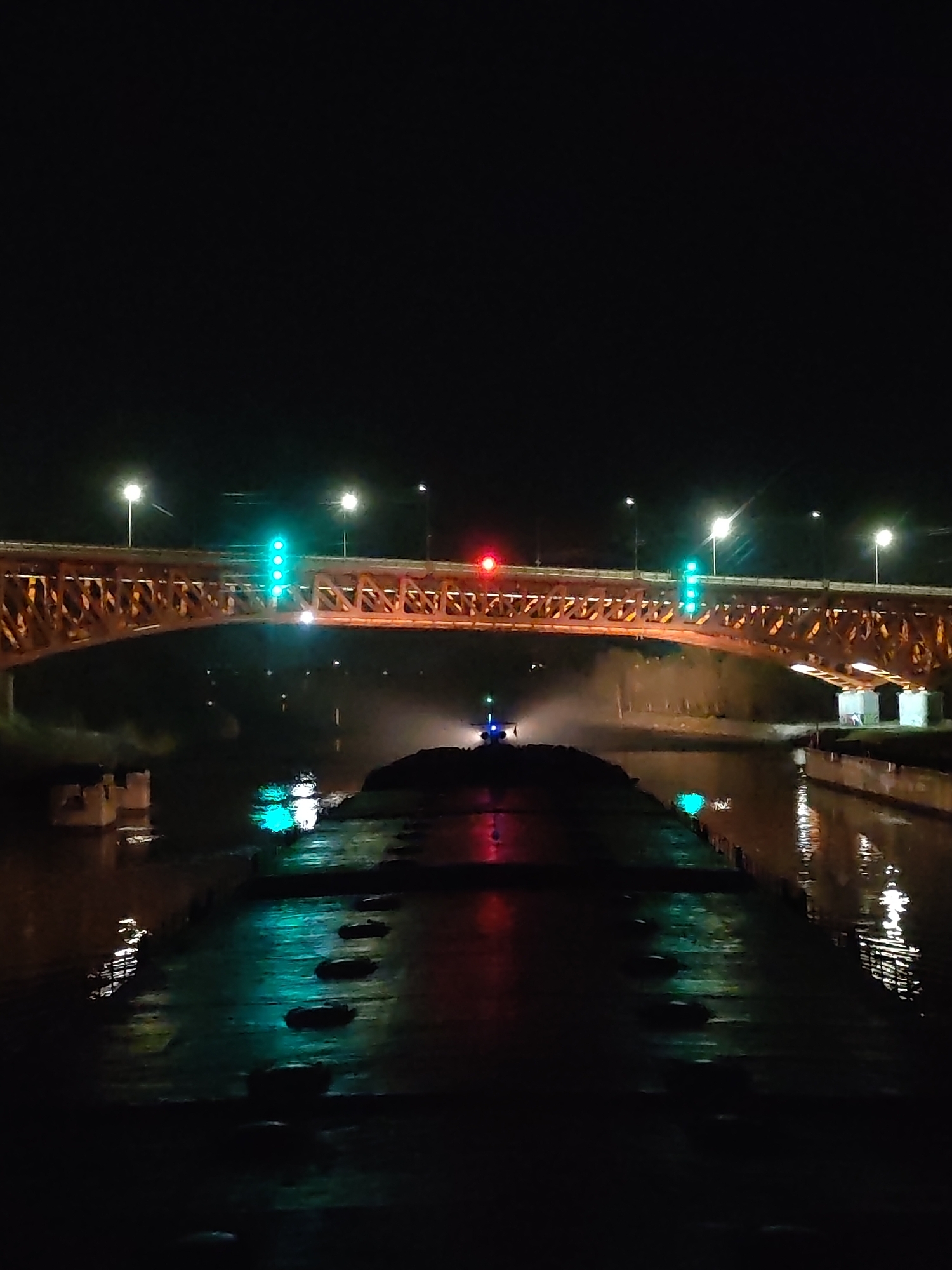 The image depicts a nighttime scene of a river and bridge. The bridge has a lattice structure and is illuminated by streetlights, with what appears to be green and red traffic signals. Below the bridge, a vessel is visible on the river, with a green light at the front and red light at the rear. The water reflects these lights, and the vessel appears to be a large barge, potentially composed of multiple sections. The surrounding environment is dark, with only the artificial lights visible. A building structure is visible on the right side of the bridge.