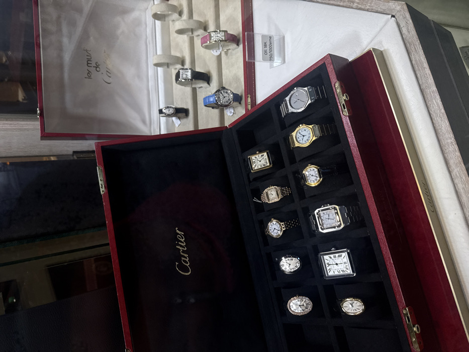 The image displays a luxury watch retail showcase, likely situated within a Cartier boutique or an authorized reseller in Paris, France. In the foreground, a large, open, deep red presentation box, lined with black velvet and featuring the "Cartier" logotype in gold on its inner lid, contains ten Cartier wristwatches. These watches are arranged in individual, black-lined compartments and exhibit various styles, including rectangular, oval, and round cases, with a combination of metallic bracelets and leather straps, presented in gold and silver-toned finishes. To the right and slightly behind this primary box, a second, open red box, similarly styled but with a white satin-lined inner lid bearing the "les must de Cartier" logotype in gold, serves as a backdrop. Below this second box, on a white display surface, five additional Cartier watches are individually presented on small, light-colored stands, alongside several empty matching stands. A clear acrylic sign positioned near this secondary display reads "OR 18K 750/1000eme", indicating 18-karat gold content. The scene is brightly lit, focusing exclusively on the displayed timepieces; no individuals or active interactions are visible.