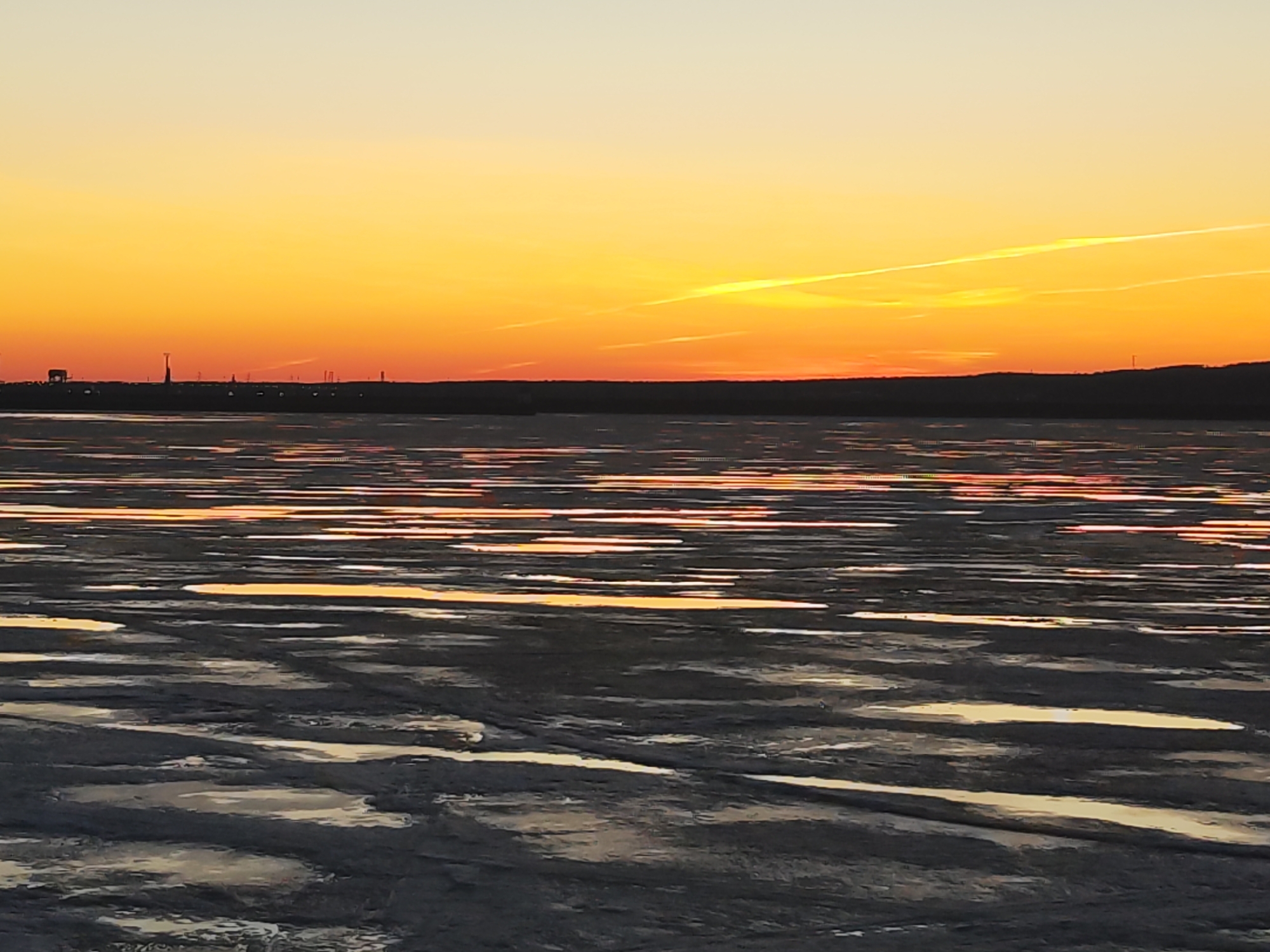 The image depicts a partially frozen body of water at sunset in Chaikovsky, Russia. The sky transitions from pale yellow near the horizon to a deeper orange hue higher up, with contrails visible. The water's surface is a mixture of ice and open water, with the setting sun reflecting off both surfaces. The land in the distance appears as a dark silhouette, with a few distinct structures visible on the horizon. The foreground shows the icy surface of the water, which is gray with patches of open water reflecting the sunset colors. The scene is quiet and still, suggesting a cold or possibly frozen environment.