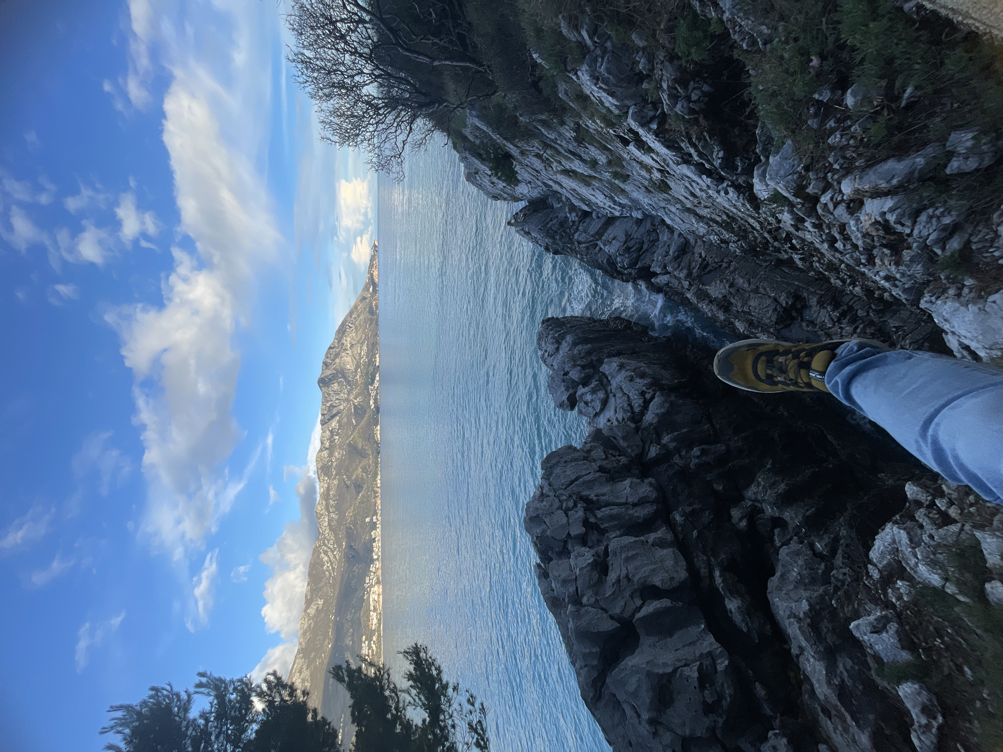The image displays a coastal landscape in Budva, Montenegro, viewed from a high rocky outcrop. In the immediate foreground, a person's lower right leg is visible, clad in blue denim jeans and a yellow/tan hiking boot, extended horizontally over the precipitous dark grey rock face. The rock formations feature jagged edges and crevices, descending steeply towards the sea where waves are observed breaking. The midground consists of the calm, blue-green water of the Adriatic Sea. Across the bay, distant rugged mountains form the background, their lower slopes featuring numerous buildings indicating a coastal town or settlement, consistent with the Budva area. The sky above is blue with scattered white cumulus clouds. Sparse green vegetation, including a dark pine tree on the left and a bare, branched tree on the right, frames parts of the scene.