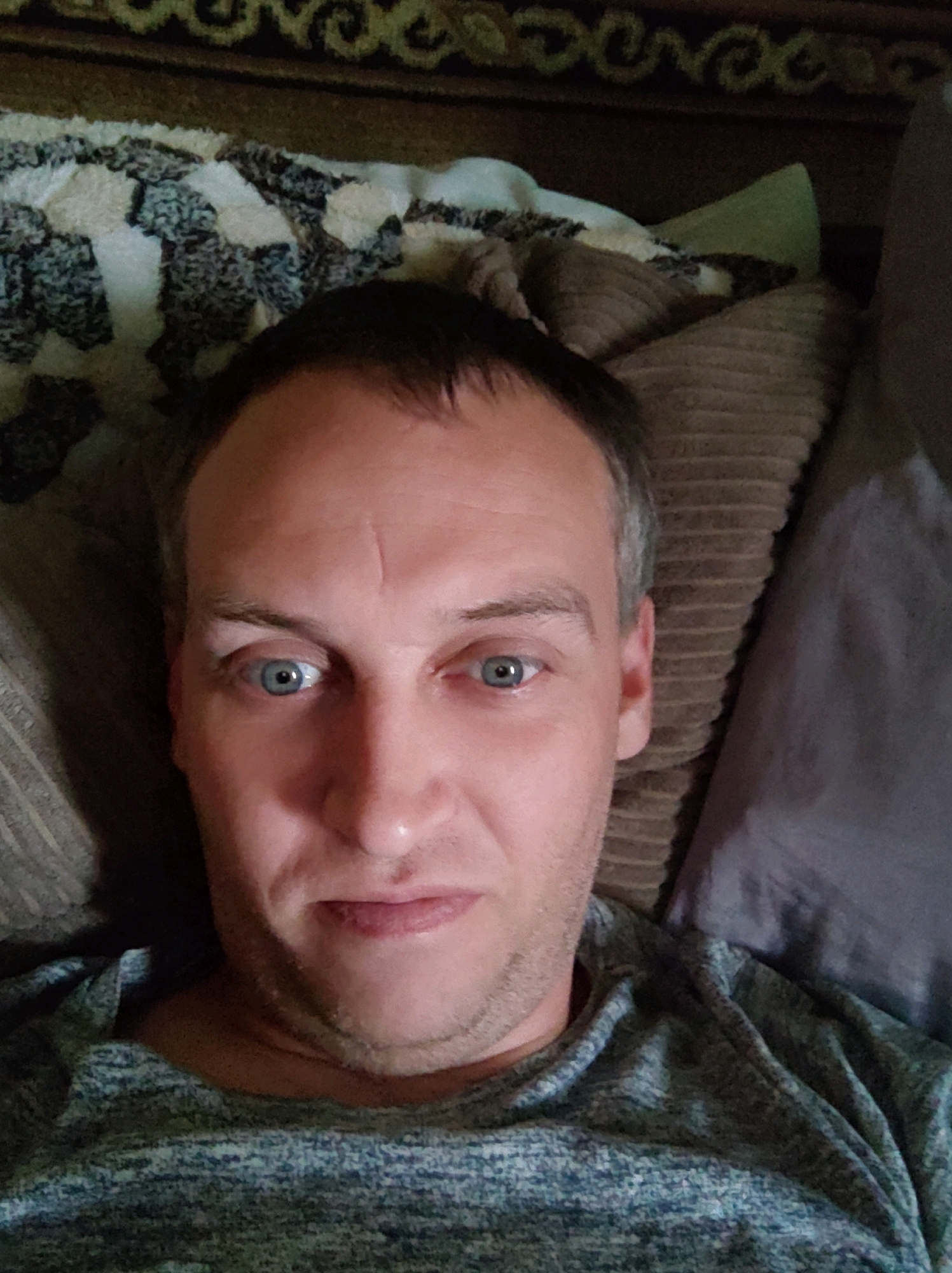 A close-up, overhead, selfie-style photograph shows a fair-skinned man lying down, with his head resting on a pillow. He has short, dark brown hair, a fair complexion, and blue eyes. He is wearing a grey, speckled t-shirt. The background consists of a patterned blanket and pillows, suggesting an indoor setting. The man is the sole subject of the photograph. The image does not contain any information about the location in the city of Stryhantsi, Ukraine.