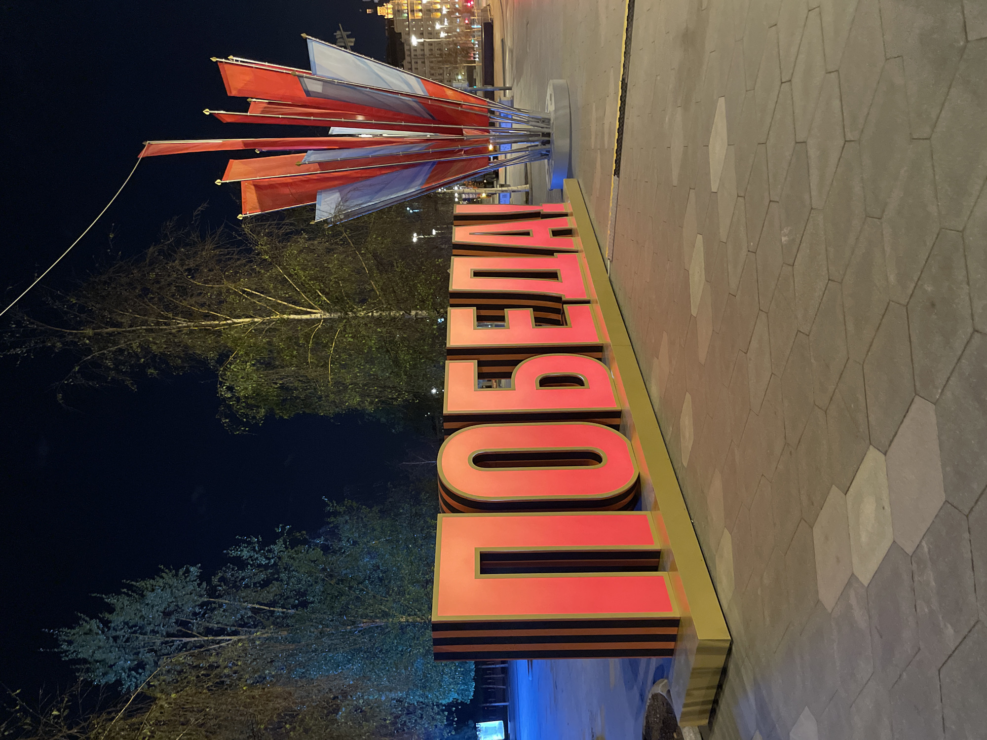 The image is a night-time shot of a public space, likely in Moscow, Russia, featuring a large illuminated sign in Cyrillic script, "ПОБЕДА!" (Victory!). The sign is constructed of red letters with a black and orange striped base, reminiscent of the Ribbon of Saint George, a Russian military symbol. Behind the sign are several bare trees, and a pole with multiple red and blue flags. In the background, there are some blurry buildings with lights on, giving a sense of a city at night. The foreground features a paved area with grey hexagonal stones. The photo is taken at ground level and the scene is somewhat sideways. There are no people visible in the shot.