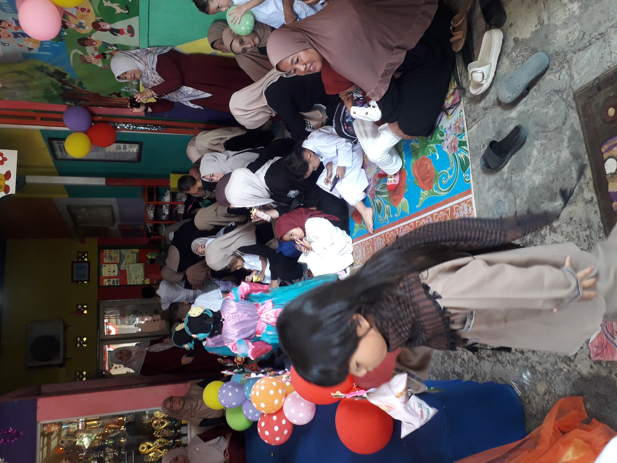 In Cankuang, Indonesia, a group of people, predominantly women and children, are gathered indoors for what appears to be a cultural or festive event, possibly a celebration or a school activity. The room is brightly decorated with colorful balloons and murals. A woman in a maroon hijab and dress is photographing the scene with a smartphone. Children in traditional attire, including one in a pink and blue dress, are among the attendees. Many of the adults are wearing hijabs. The floor is covered with a patterned rug, and various footwear, including sandals and slippers, are scattered around. The environment suggests a community or educational setting.