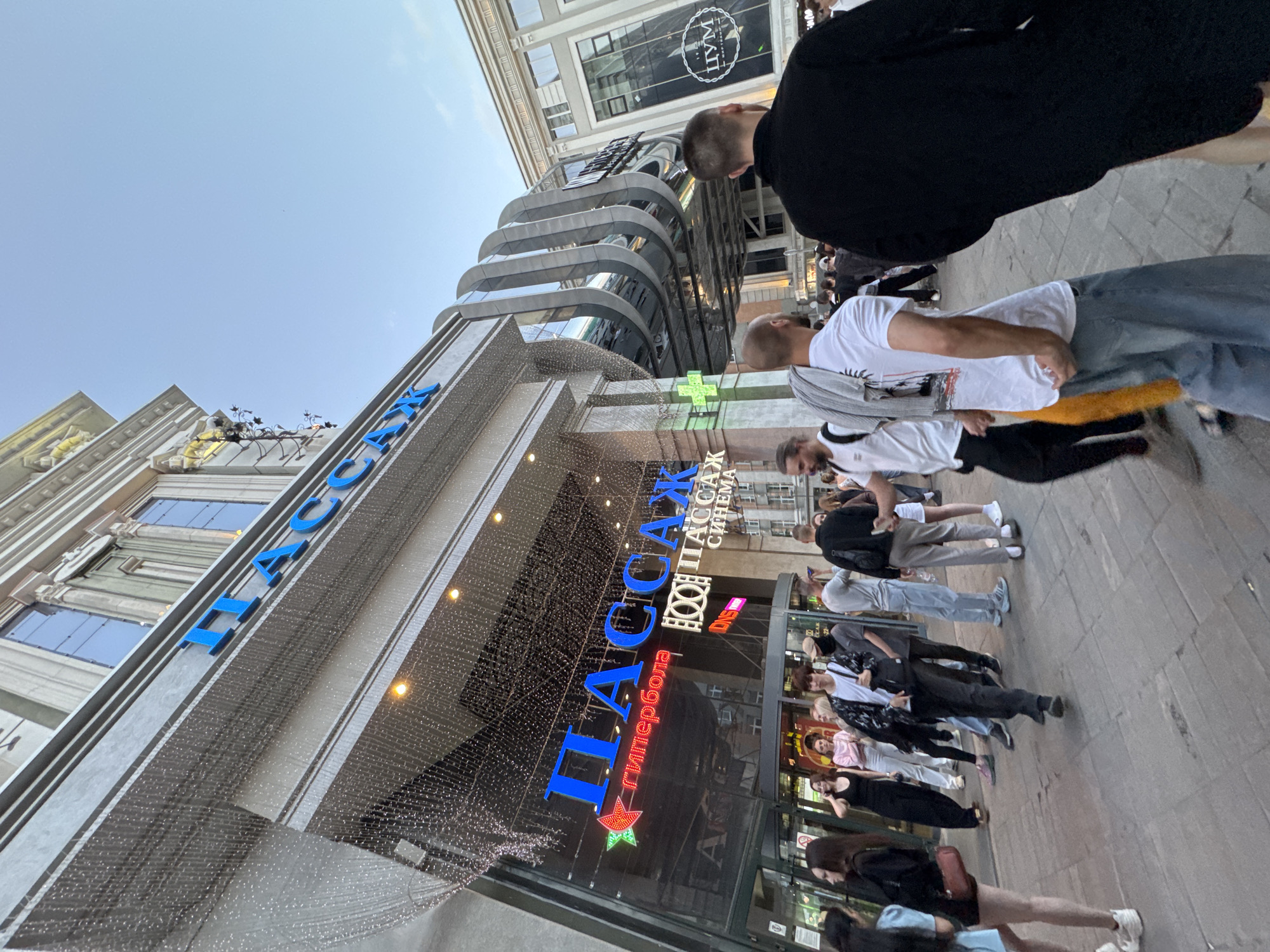 The image shows the entrance to a shopping center in Yekaterinburg, Russia. The shopping center is named "PASSAGE". The entrance is large with glass doors and many people are walking in and out. Above the entrance, there are large blue letters spelling out "PASSAGE" and also "PASSAGE CINEMA". To the left of the entrance, there is a neon sign with a red star and the word "ГИПЕРБОЛА" (hyperbola). The architecture of the building is modern with decorative elements and lights. Several people are visible in the foreground and midground, dressed in casual clothing. The sky is clear and blue. The overall scene depicts a busy urban environment outside a shopping mall.