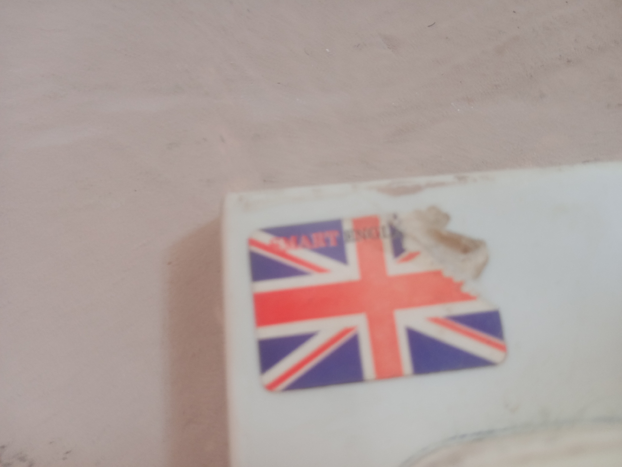 The image displays a close-up, blurry view of a sticker featuring the Union Jack flag, overlaid with text that appears to read "SMART ENGL". A torn piece of what looks like a receipt or label is attached to the top right of the sticker. The sticker is affixed to a white surface, possibly a book or a box, which is set against a textured, light pinkish-beige background, likely a wall. The overall scene is underexposed and lacks clear detail due to the focus and lighting. The image is not specific enough to determine its location in Jalingo, Nigeria.