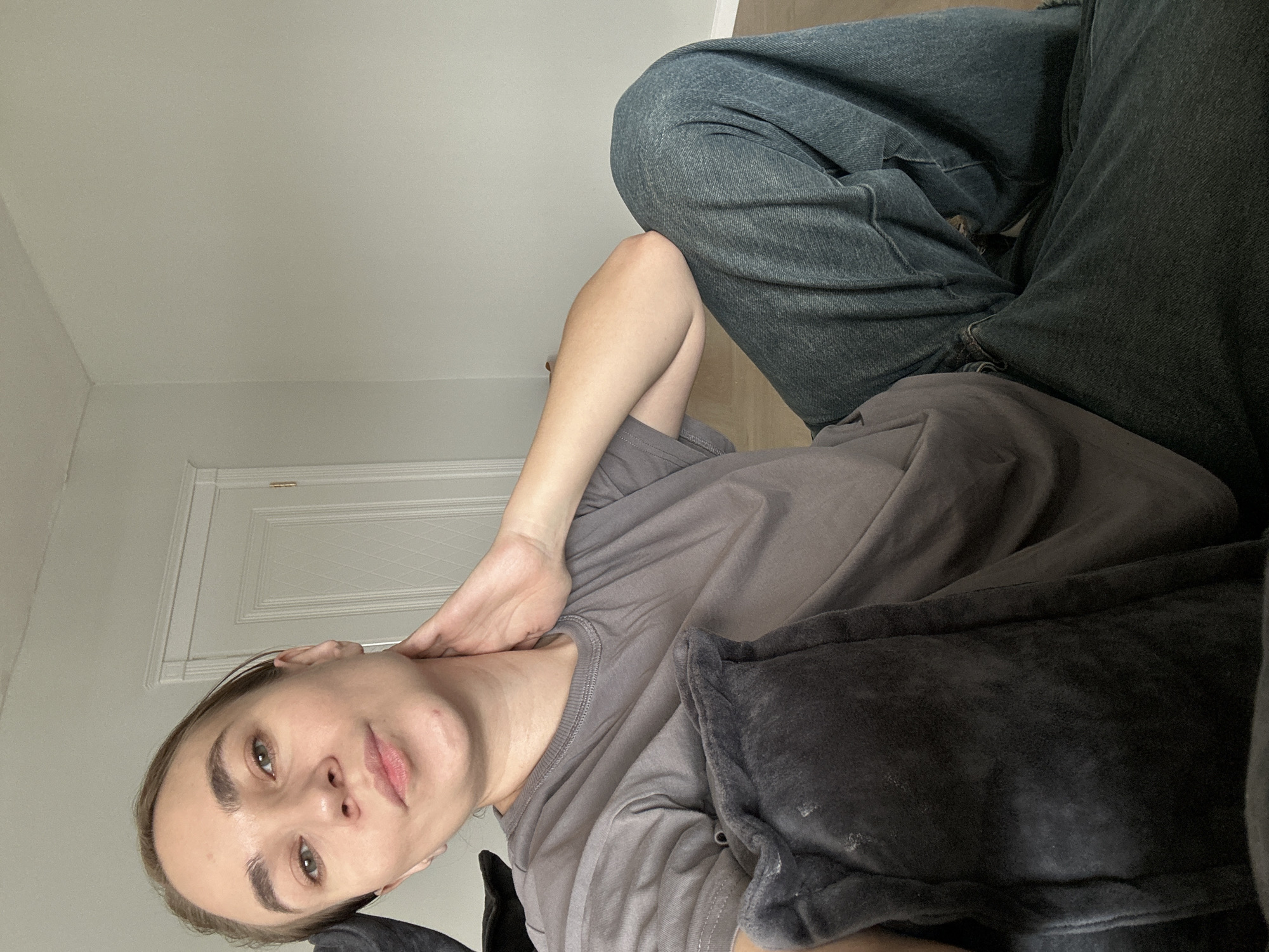 A woman is reclining on a soft surface, possibly a couch or bed, wearing a grey t-shirt and denim jeans. Her head is resting on a dark grey pillow, and her right hand is touching her neck. The background features a white wall and a closed white door. There is no evidence to confirm the location is Kharnavy, Russia.