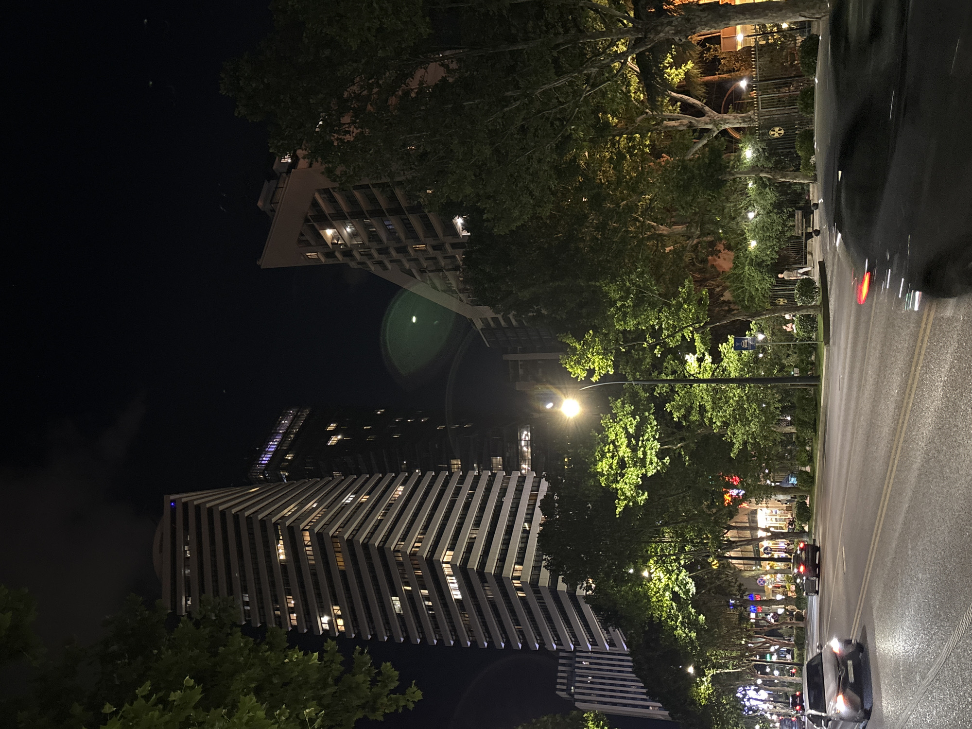 The image captures a night scene in a city, featuring modern architecture and urban infrastructure. A prominent, tall, striped building with numerous illuminated windows stands on the left, partially obscured by the branches of a large, leafy tree. To the right, another tall building with a triangular facade and balconies is visible, also with lit windows. A tree-lined street runs horizontally across the foreground, with streetlights casting a warm glow on the pavement and foliage. Several cars are present on the road, with one silver car in the left lane and a dark car in the right lane. The right side of the image shows a black car in motion, with its taillight creating a red streak. Beyond the trees, a fence with decorative metalwork and some illuminated structures can be seen. The sky above is dark, indicating nighttime, with faint lights and possibly some clouds. The overall impression is that of a bustling city at night, with a mix of residential and commercial buildings.