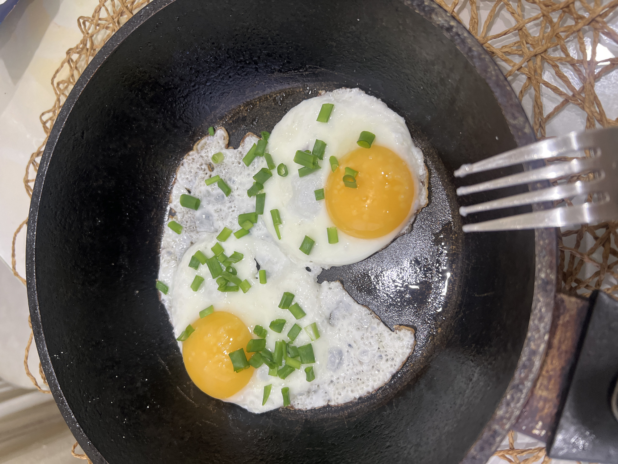 A close-up shot depicts a black cast-iron frying pan containing two fried eggs, sprinkled with chopped green onions. A silver fork is positioned near the right edge of the pan. The pan rests on a woven placemat. The location is not identifiable from the image.