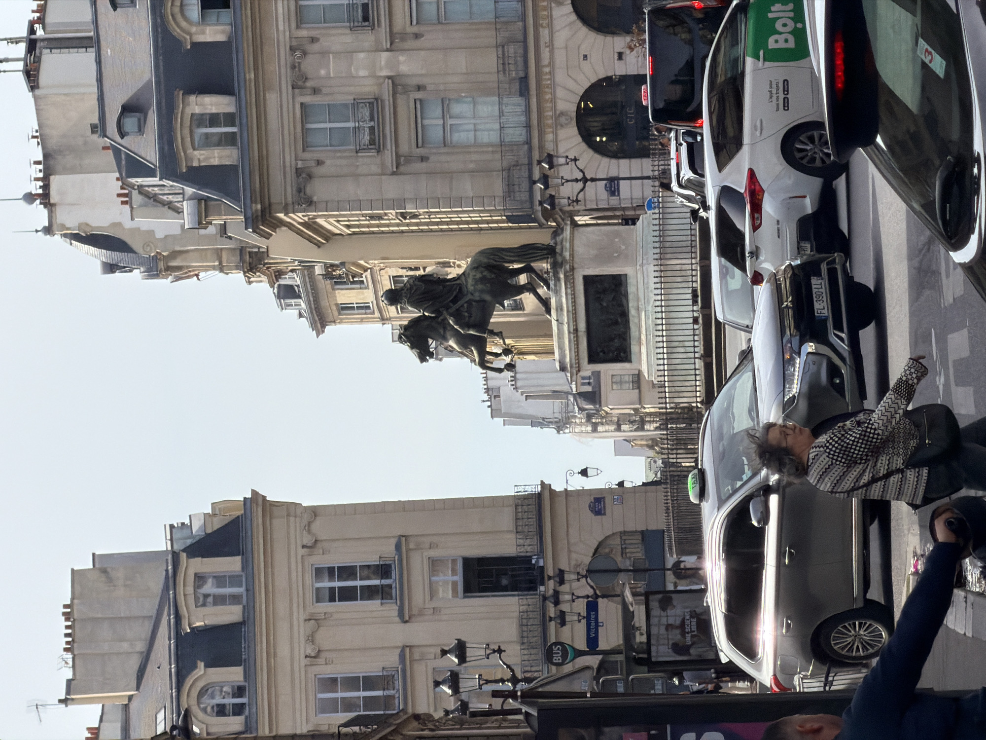 An upright, daylight street view in Paris, France, captures a bustling urban scene with historic architecture and modern traffic. On the left, a row of ornate, light-colored Parisian buildings with multiple windows and detailed facades lines the street. In front of these buildings stands a large, dark bronze equestrian statue on a stone pedestal, depicting a historical figure on horseback, with additional sculptural elements at its base.
The right side of the frame is dominated by a street with several parked or moving cars. A prominent white Bolt ride-share car with green branding is visible, displaying the text "Bolt" and "L'appli pour tous vos trajets!" (The app for all your trips!). Closer to the viewer is a silver SUV, its side reflecting bright sunlight. A woman with grey, curly hair, wearing a black and white patterned top and carrying a dark shoulder bag, is visible near this SUV, appearing to be walking or standing on the sidewalk. Part of another person's arm and shoulder is visible in the extreme foreground.
Visible text and signs include "BUS" and "Victoires" on a street sign or bus stop, indicating the location is likely near Place des Victoires. Other text includes "CIC" on a building or sign, "FL-390-LL" on a French license plate, and "UNE SCIENCE LIBRE" on a poster or advertisement. A small red "3" is also seen on the rear window of a car. The bright sunlight suggests clear weather during the day.