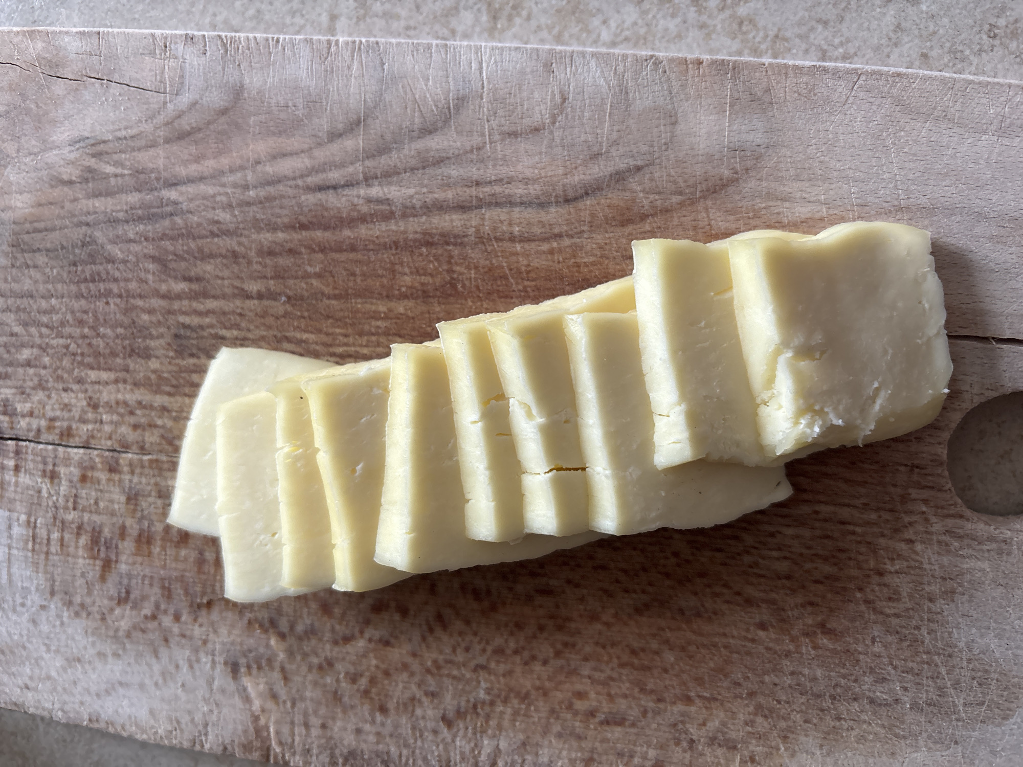 A pile of sliced cheese is arranged diagonally on a wooden cutting board. The cheese is pale yellow and appears to be a firm variety. The cutting board has visible grain and some scratches. The background is out of focus and appears to be a countertop. There are no people or animals in the image. This image was captured in Bila Tserkva, Ukraine.