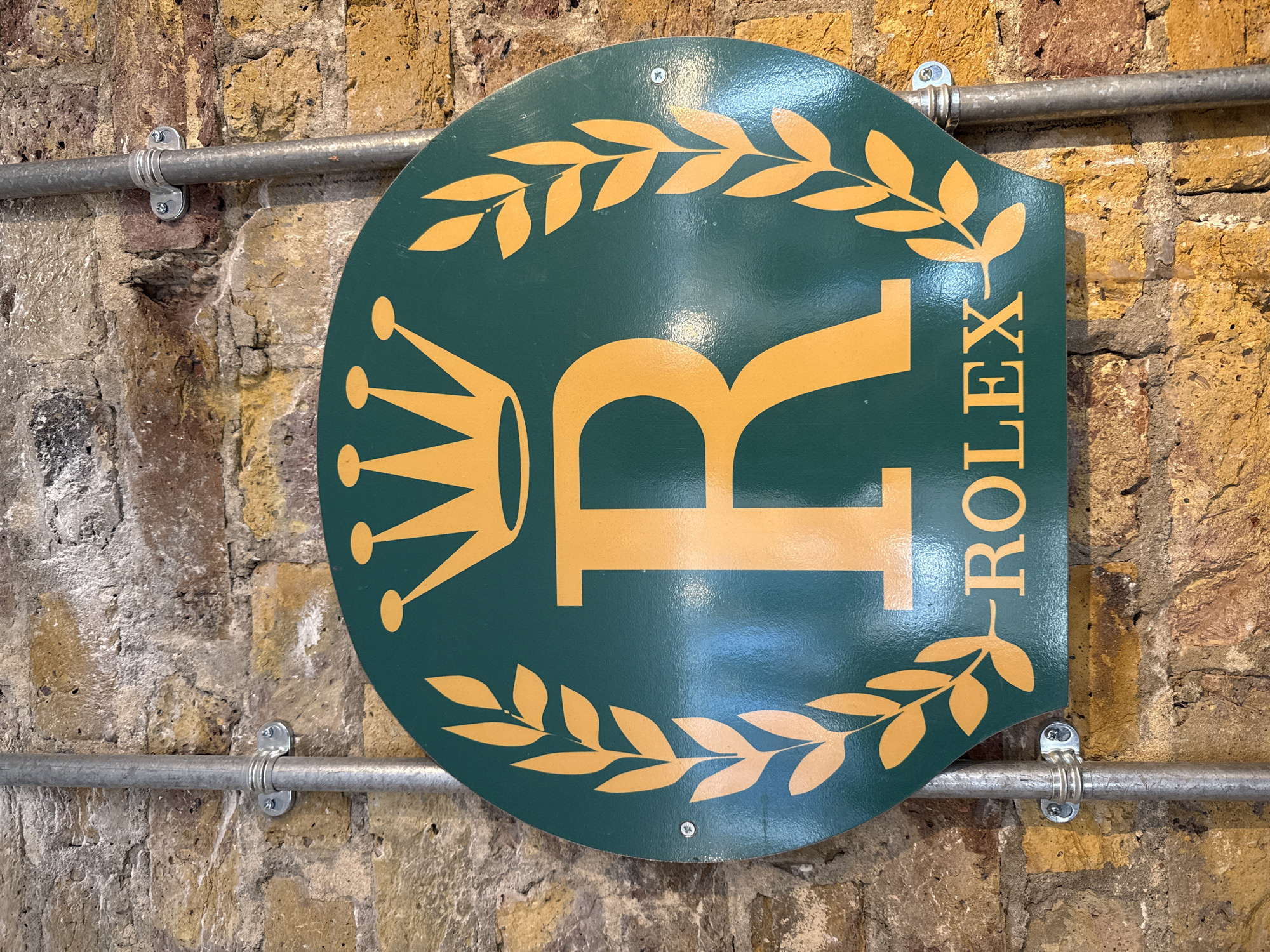 The image depicts a Rolex sign mounted on a brick wall. The sign is circular with a flat top. It's dark green with a gold-colored laurel wreath design around the top and bottom. A large gold-colored letter "R" is prominently displayed in the center, with a gold-colored crown to the left and the word "ROLEX" vertically aligned to the right of the "R". The sign is attached to a brick wall with two silver-colored metal pipes running horizontally across it. The pipes are fixed to the wall with brackets. The brick wall appears aged and textured. The location is implied as London, United Kingdom due to the Rolex sign and typical brickwork seen in the city.