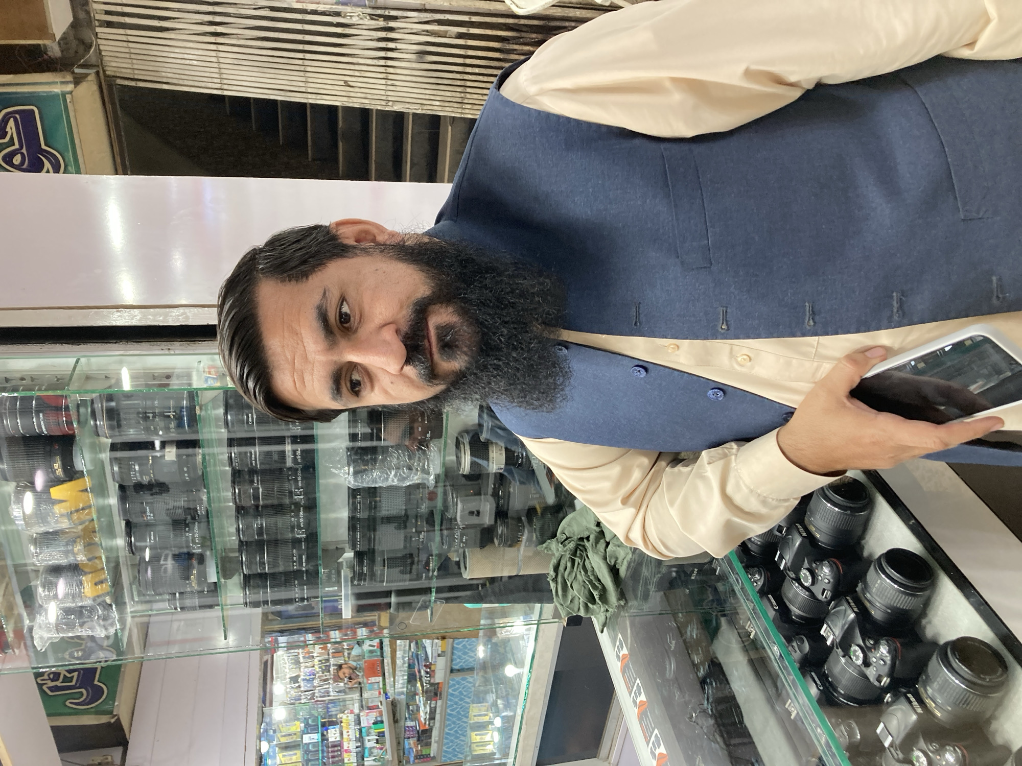 A middle-aged man with a beard stands in front of a glass display case filled with cameras and lenses in Quetta City Tehsil, Pakistan. He wears a light-colored shirt and a navy blue vest and holds a smartphone in his right hand. The display case contains multiple cameras and various lenses, and reflections can be seen in the glass. A store sign is visible above the display case. The area behind the display case appears to be a shop with shelves and products.