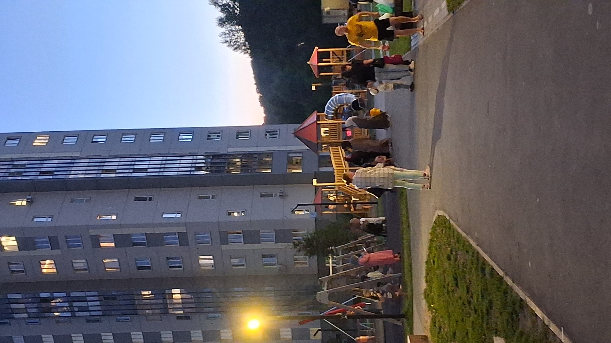 The image shows a multi-story apartment building with many windows, some of which are illuminated. In front of the building is a playground with several structures, including a slide and swings. Several people, including adults and children, are visible on the playground and in the surrounding area. The scene appears to be a public space in a residential area. The lighting suggests it is either early morning or late evening. There is a streetlamp on the left side of the image. A hilly, wooded area is visible in the background, beyond the apartment building. This scene is a common sight in residential areas of cities like Novosibirsk, Russia, especially in the evening when families often gather at playgrounds.