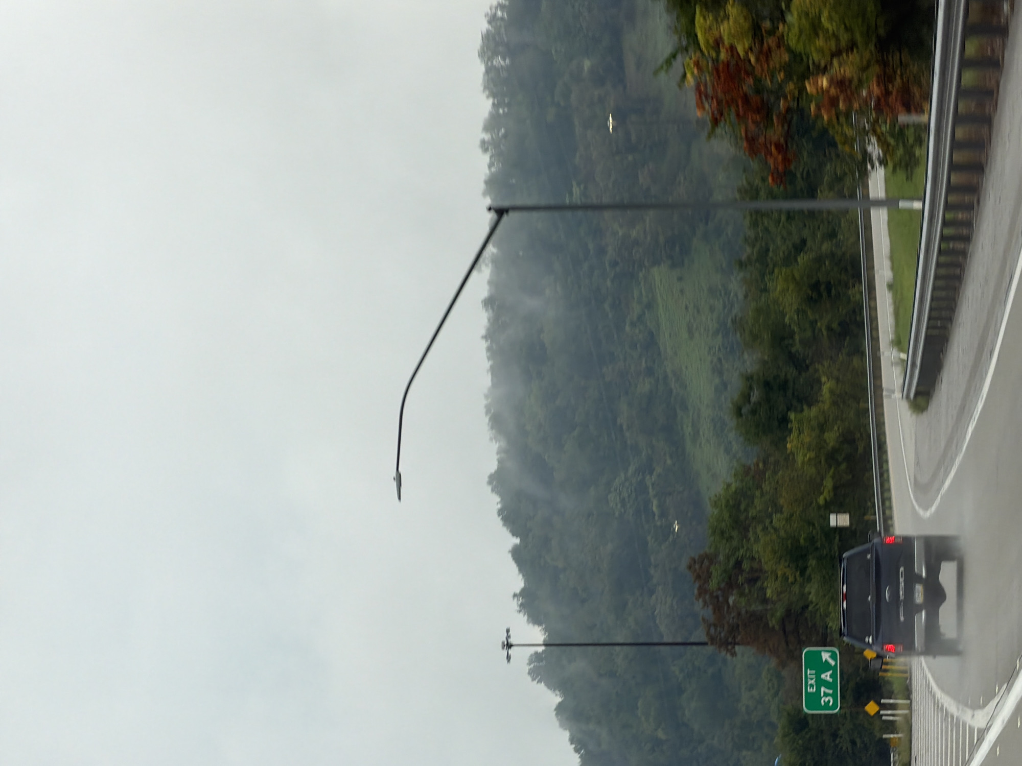 A dark-colored Ford F-150 pickup truck is driving on a wet road in Fallowfield Township, USA. The truck is in the foreground, driving away from the camera, with its taillights illuminated. The road curves to the right and is wet, with visible spray coming from the truck's tires, indicating recent rain or damp conditions. To the left of the road is a green highway sign indicating "EXIT 37 A" with an arrow pointing to the right. There are also yellow diamond-shaped signs nearby. In the background, there is a lush green hillside covered in trees, with some fog or mist visible amongst the trees. A tall light pole with multiple fixtures stands on the left side of the road. A curved lamppost is also visible in the upper right quadrant of the image. The sky is overcast and gray.