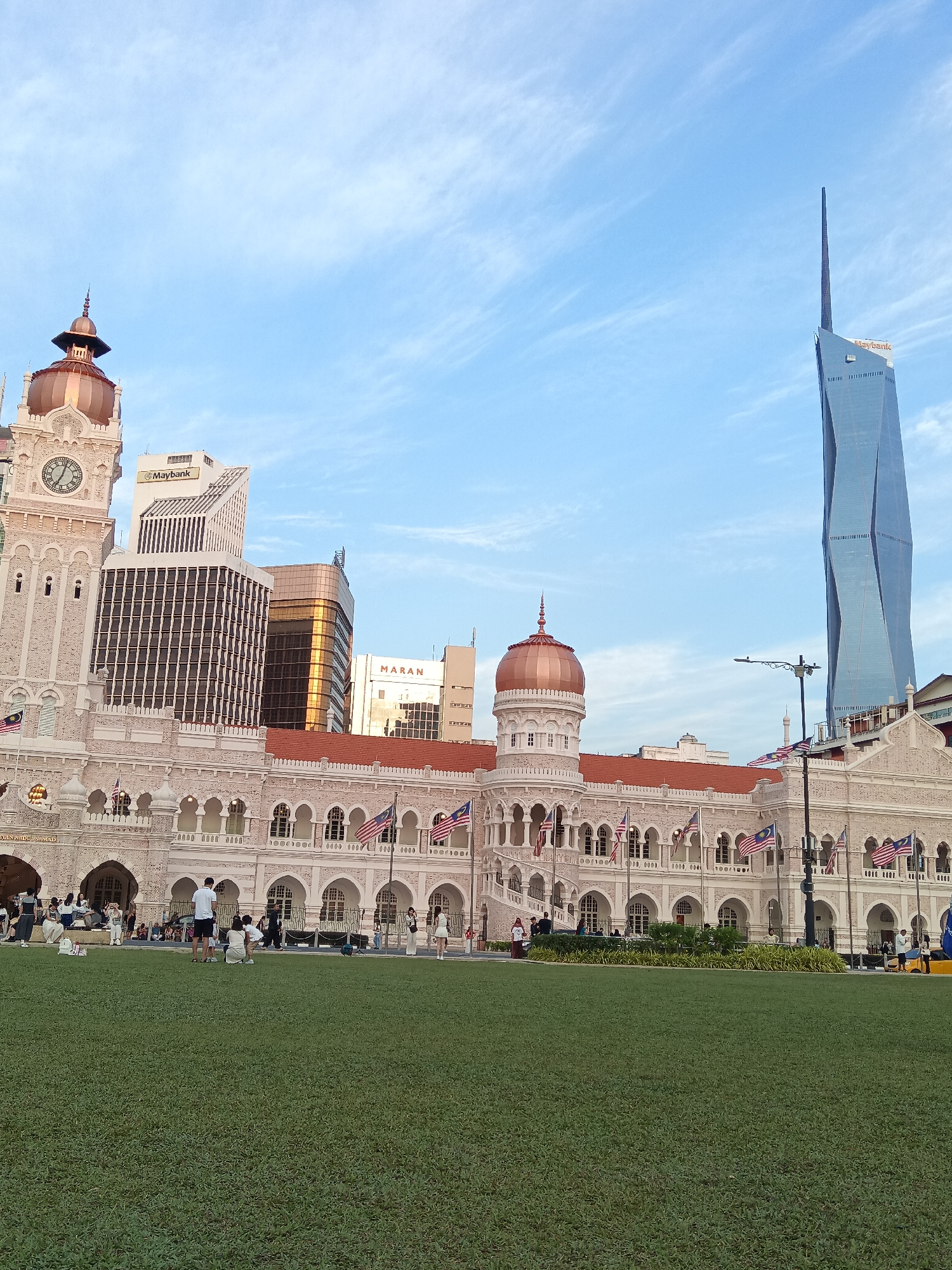 The image depicts the historic Sultan Abdul Samad Building in Kuala Lumpur, Malaysia, situated in front of a lush green field with several individuals on it. The building, characterized by its distinctive copper domes and intricate arches, stands prominently in the foreground, adorned with numerous Malaysian flags. Behind the Sultan Abdul Samad Building, a cluster of modern skyscrapers rises, including the distinctive twisting form of the Ilham Tower. The sky above is a clear blue with wispy white clouds. The scene appears to be set during daylight, possibly late afternoon, given the soft lighting.