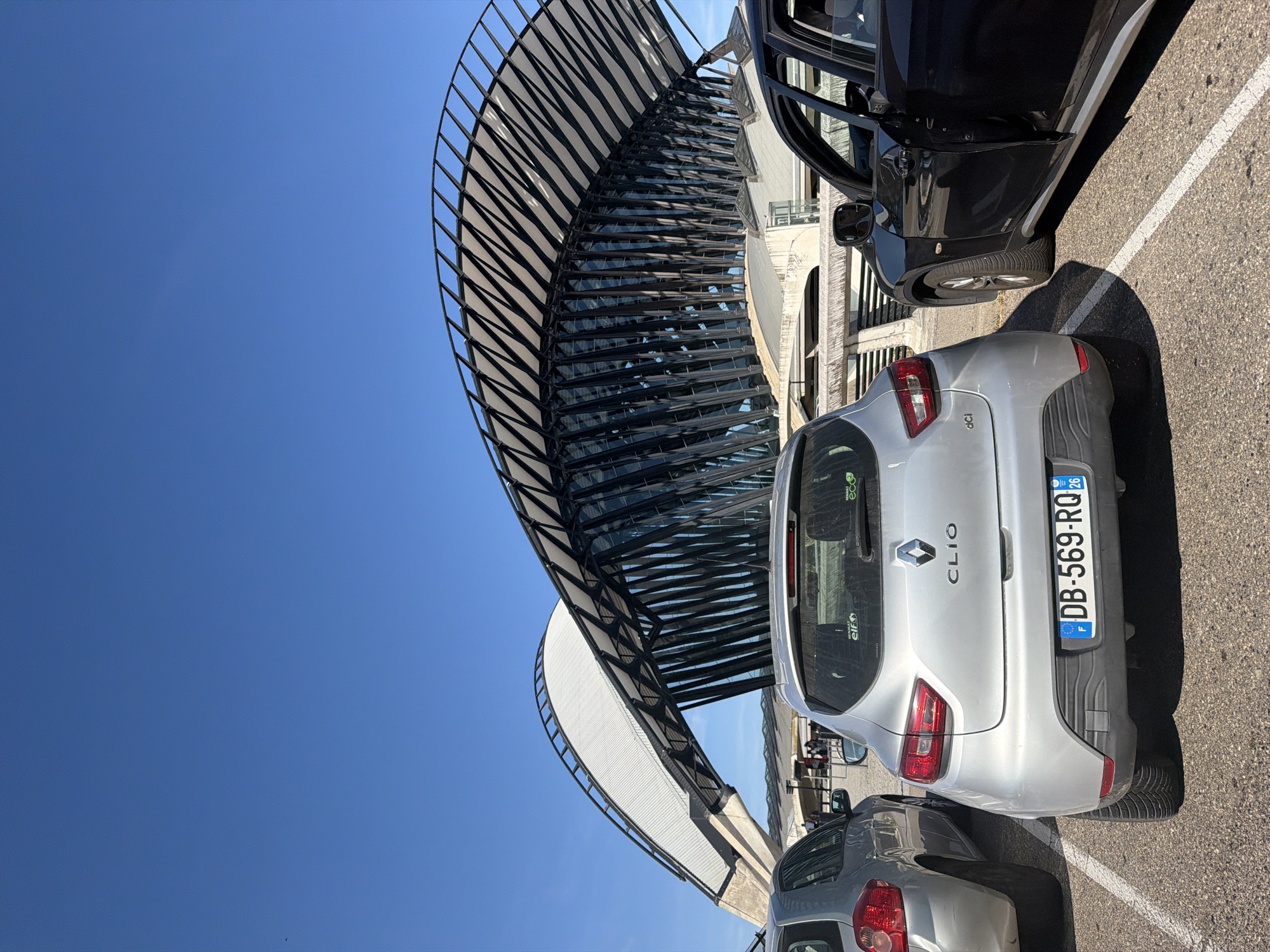 The image shows a street view with a distinctive modern building in the background. The building features a large, curved, white roof supported by an intricate black metal structure, resembling a wave or a bird's wing. In the foreground, two cars are parked on asphalt. The car on the left is silver and partially visible, while the car in the center is a silver Renault Clio with a visible license plate that reads "DB-569-RQ". To the right, a black car with its driver's door open is parked. The sky is clear and blue. This location is the Lyon–Saint-Exupéry Airport (LYS) in Colombier-Saugnieu, France, with the building being its iconic airport station architecture.