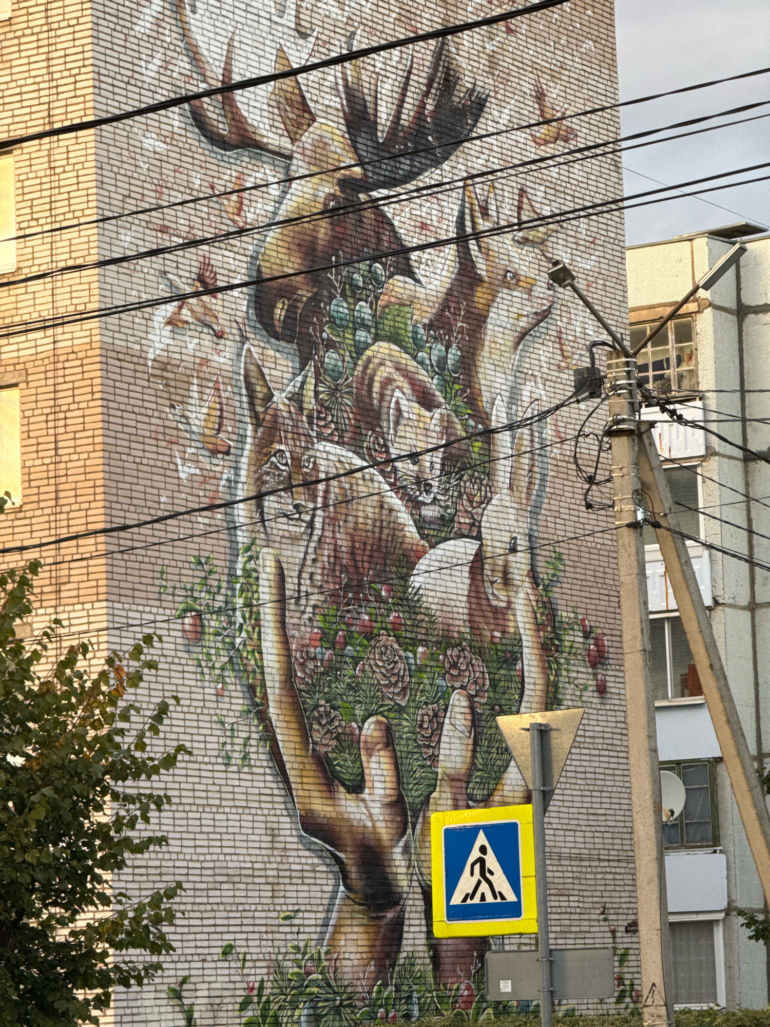 A mural depicts a collection of woodland animals, including a moose, deer, fox, lynx, and rabbit, intertwined with flora such as roses, pinecones, and berries. The artwork is painted on a brick building. In the foreground, a pedestrian crossing sign with a blue and yellow square sign featuring a white pedestrian figure is mounted on a metal pole. Electrical wires are visible in the frame, running horizontally across the mural and towards a concrete utility pole. A green tree branch with leaves extends from the left side of the image. The scene appears to be situated in the city of Valday, Russia, based on context.