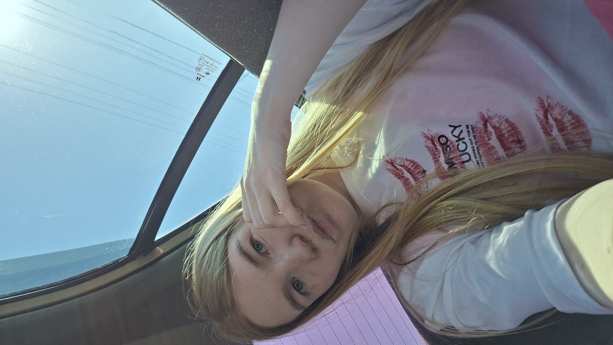A selfie, possibly taken inside a car, shows a young woman with long blonde hair, wearing a white t-shirt printed with red lipstick marks and the text "SO LUCKY." Her left hand is near her face. The background shows a car windshield with a blue sky visible through it.