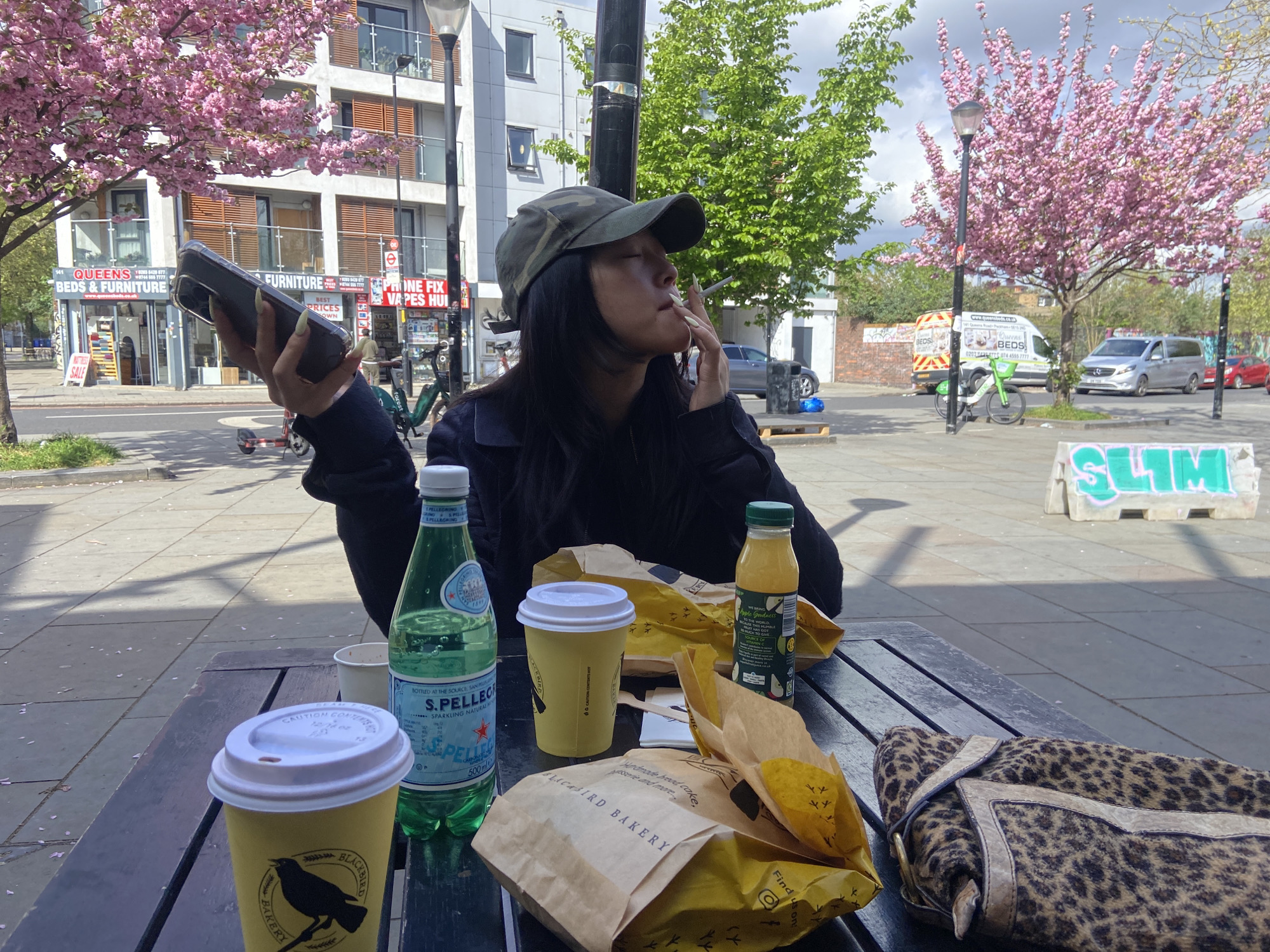 A woman sits at a picnic table outdoors in London, UK. She wears a camo cap and black jacket, smoking a cigarette and holding a phone. The table holds two cups from Blackbird Bakery, a bottle of S.Pellegrino, an unlabeled bottle of juice, and several paper bags with food. A leopard print purse is on the table's edge. Across the street are shops including Queens Beds & Furniture and Phone Fix Vapes Hui. Cherry blossom trees are in bloom. A van and parked cars are visible in the background. A green Lime e-bike stands nearby.