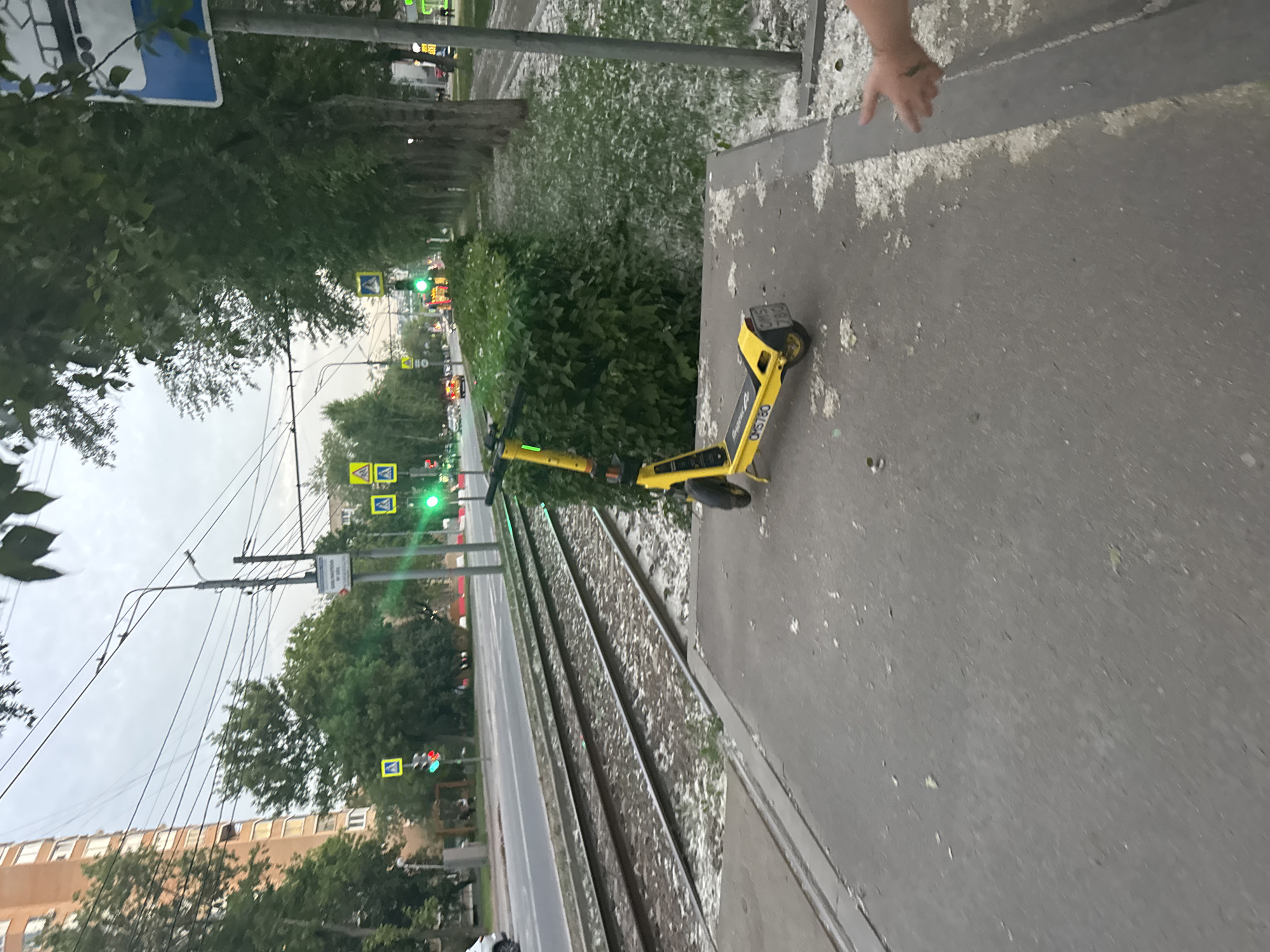 An outdoor urban scene depicts a sidewalk next to tram tracks and a road. A yellow electric scooter is parked on the sidewalk. Across the road, traffic lights and pedestrian crossing signs are visible. Trees line the side of the road opposite the tram tracks, with a building in the background. The ground on the sidewalk and the tram tracks are littered with white debris, possibly snow or ice. A person's hand is partially visible in the lower right corner of the image, reaching towards the debris.