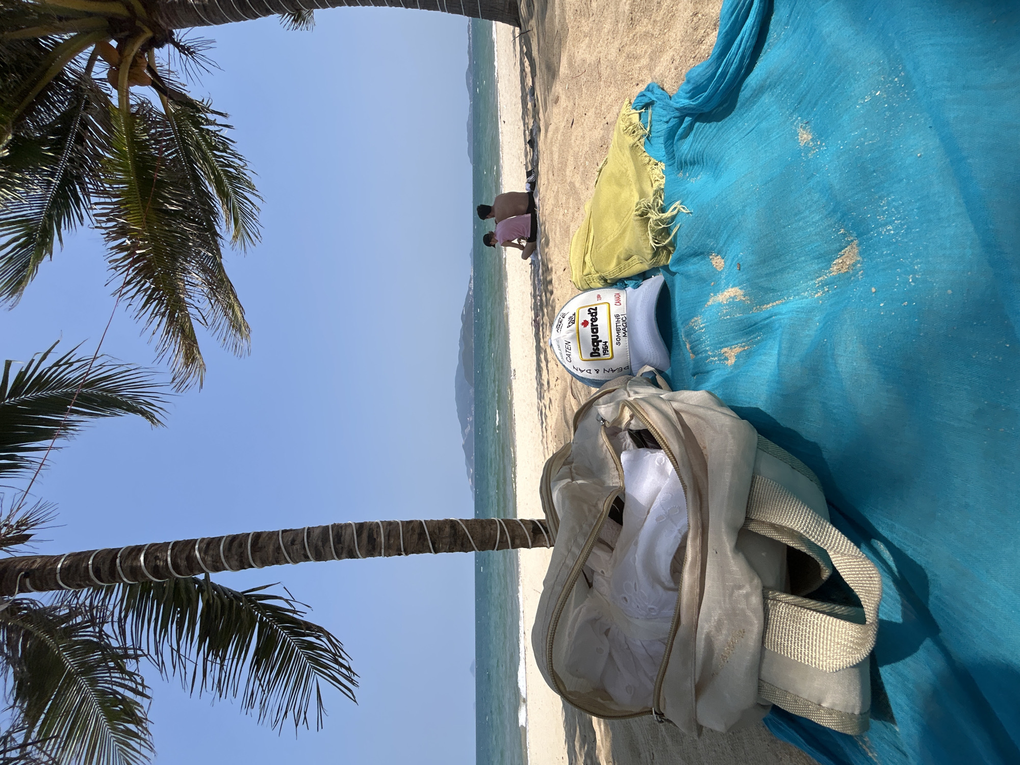 The image shows a beach scene in Nha Trang, Vietnam. The foreground features a blue and yellow patterned blanket, a partially open off-white backpack, and a white baseball cap with black and red lettering. Two palm trees frame the left side of the image, their fronds extending into the blue sky. In the background, two individuals are sitting on the sand near the water's edge, the sea stretching out to the horizon with distant hills.