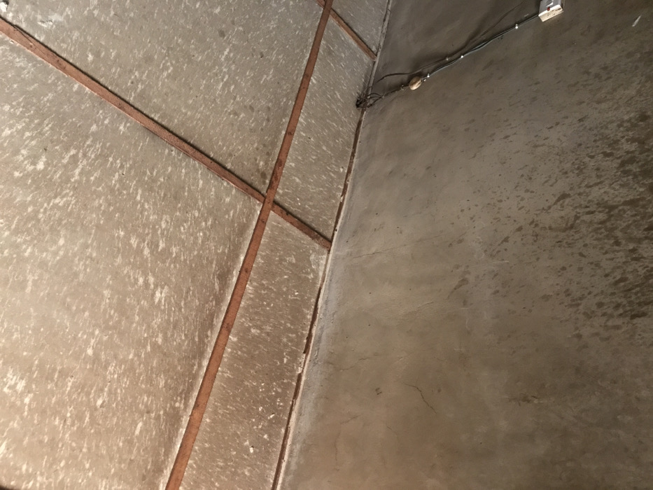 The image displays an interior corner of a room, likely a utilitarian space such as a storage area or a basement, situated in Oba-Ile, Nigeria. No people are present in the scene.
The surface on the left side of the frame consists of light grey or beige panels, possibly made of concrete or a similar material, which are separated by dark reddish-brown strips arranged in an irregular grid pattern. These panels feature a prominent mottled texture with white speckled areas, suggesting either an uneven paint job, dried water stains, or a specific textured finish.
The surface on the right is a plain, untextured wall of light grey, characteristic of concrete or plaster. Along the upper section of this wall, near the corner, black electrical wires are visibly routed. A white electrical outlet or switch is mounted on this wall, connected to the wiring, and a smaller, light-colored electrical component or fastener is also attached nearby.
The plain wall shows areas of darker discoloration or dampness, particularly towards the bottom right quadrant of the image. The overall setting appears functional rather than decorative, with visible electrical installations. There is no specific activity occurring, and no text is visible anywhere in the image. The lighting suggests an indoor environment, with no cues for the time of day or external weather conditions.