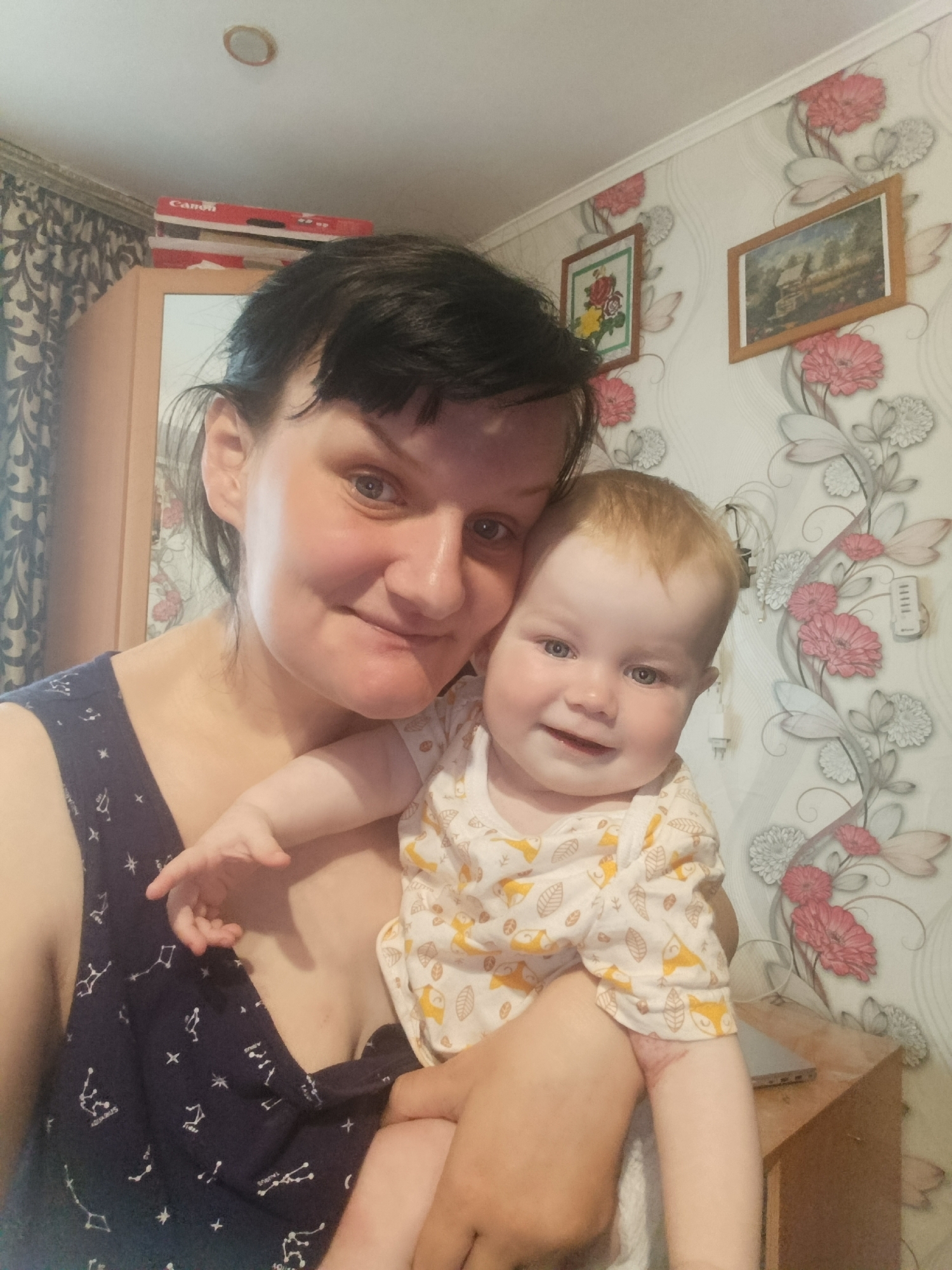 A warm and affectionate selfie captures an adult woman holding a baby in what appears to be a home interior. The woman, with dark hair, light skin, and a genuine smile, looks directly at the camera. She is wearing a dark blue sleeveless top patterned with white celestial designs, including constellations with visible text such as "Aquarius" and "Taurus."
Cradled in her arms, the baby has light blonde hair and bright blue eyes, also smiling broadly at the camera. The baby is dressed in a light-colored long-sleeved top adorned with playful yellow and brown fox and leaf patterns.
The background reveals a distinct domestic setting. The wall is covered with patterned wallpaper featuring large pink, white, and green floral and leaf motifs. Two framed pictures hang on the wall: one depicts a floral still life, and the other a landscape scene. To the left, part of a wooden cabinet or wardrobe is visible, topped with stacked red boxes, one clearly labeled "Canon." Dark, patterned curtains hang next to the cabinet. On the right side of the frame, a small wooden table holds a light-colored electronic device, possibly a laptop or tablet, and some white cables are visible against the wallpaper. A circular ceiling light fixture is also partially visible at the top. The image conveys a cheerful and loving family moment, likely taken during the daytime.