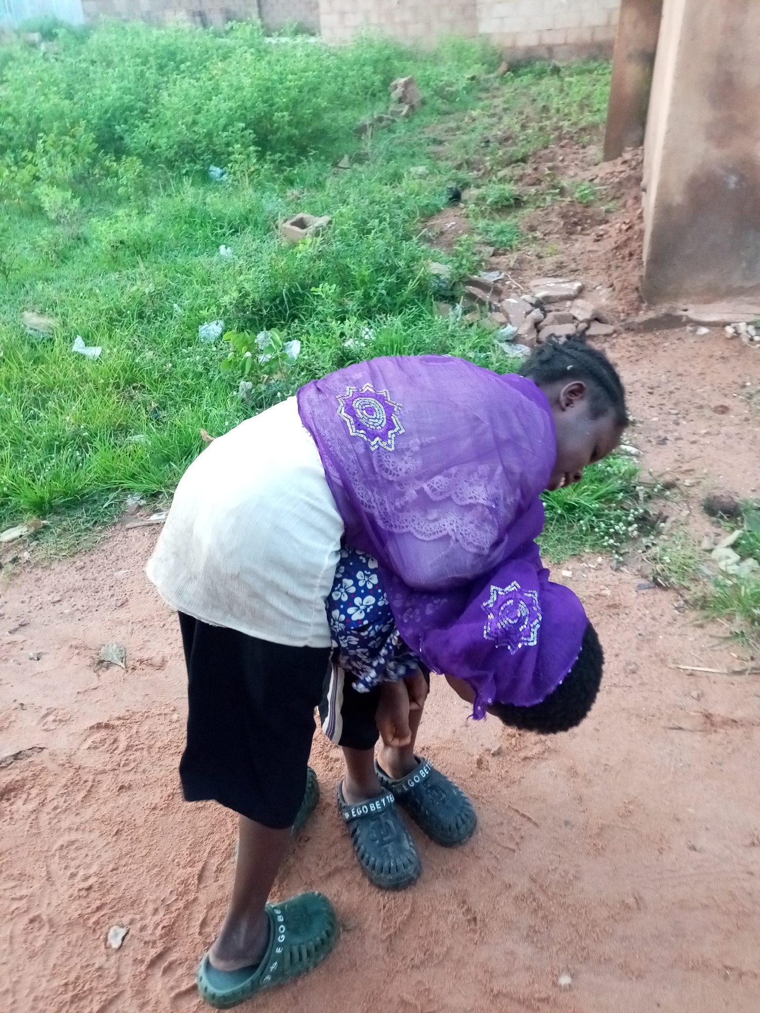 A young person wearing a white shirt, black shorts, and green clogs bends over to pick up or hold a smaller child dressed in a blue and white floral top, who is wrapped in a purple cloth. The scene takes place outdoors on a dirt path with a grassy area and a building in the background, suggesting a location in Tempure, Nigeria. The lighting indicates it is daytime.
