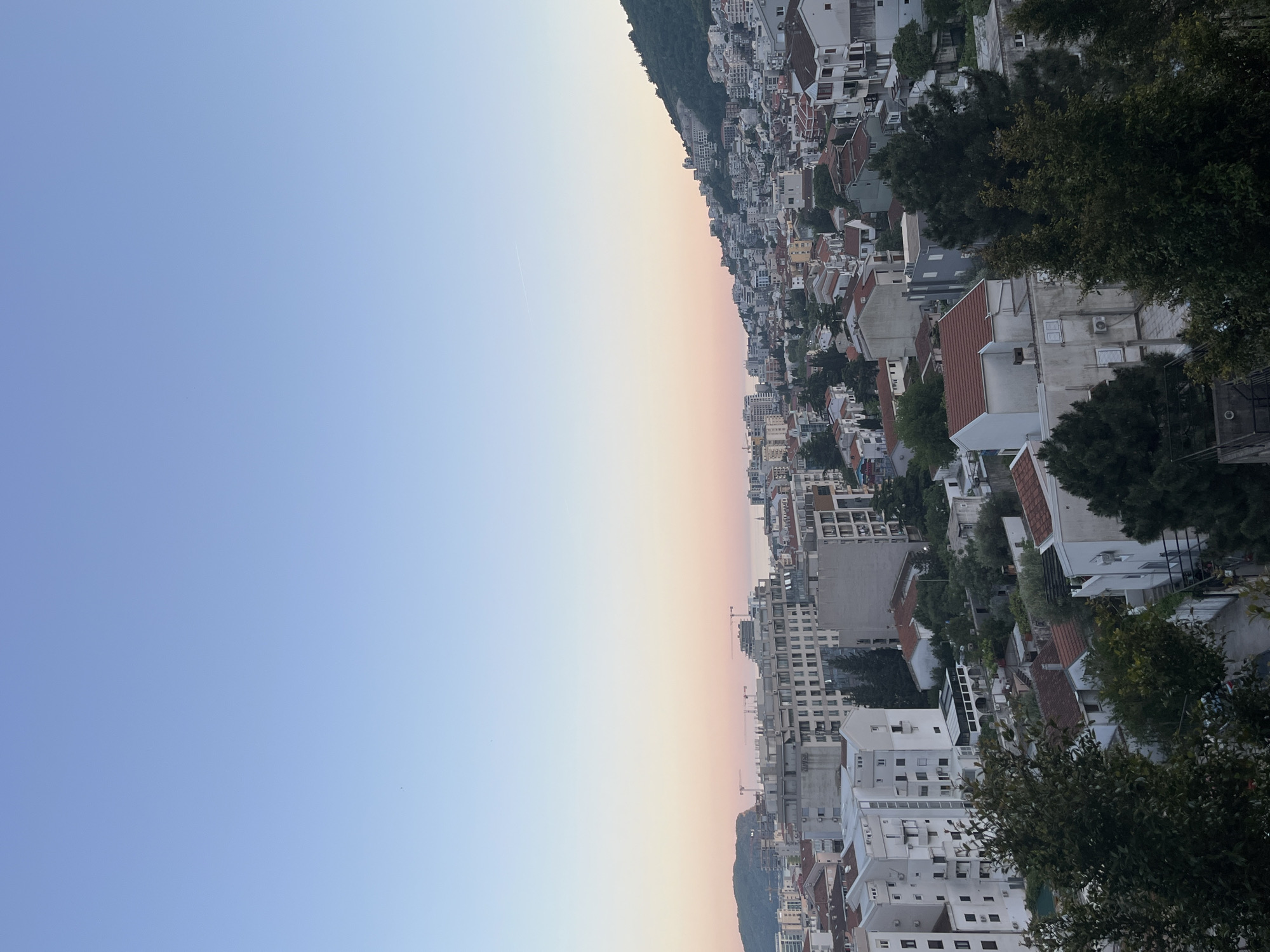 The image presents a panoramic view of a densely built coastal city or town, observed from an elevated vantage point. The scene is bisected by a vast expanse of sky above and the urban landscape below, with a faint horizon line hinting at the sea in the distance.
The sky, dominating the upper portion of the frame, transitions gracefully from a clear, light blue at the top to a soft, warm gradient of pastel pinks, oranges, and yellows towards the horizon. This coloration strongly suggests the magical hours of either sunrise or sunset. A thin, white airplane contrail is subtly visible in the blue part of the sky.
The city itself stretches across the lower half of the image, characterized by a tight cluster of buildings. Many structures feature distinct red or terracotta-tiled roofs, while others display flat, modern designs. The buildings are predominantly white or light-colored, interspersed with pockets of dark green foliage from trees and bushes, particularly in the foreground and along the hillsides. In the mid-distance, several construction cranes are visible, indicating ongoing development within the urban area.
The terrain behind the city appears to be a steep, forested hill or mountain, providing a natural backdrop to the man-made structures. The overall atmosphere conveyed by the soft lighting and clear sky is one of tranquility. No people are visible, and there is no discernible text within the image. Given the context, this scene depicts a typical coastal city in a Mediterranean region, consistent with Budva, Montenegro.