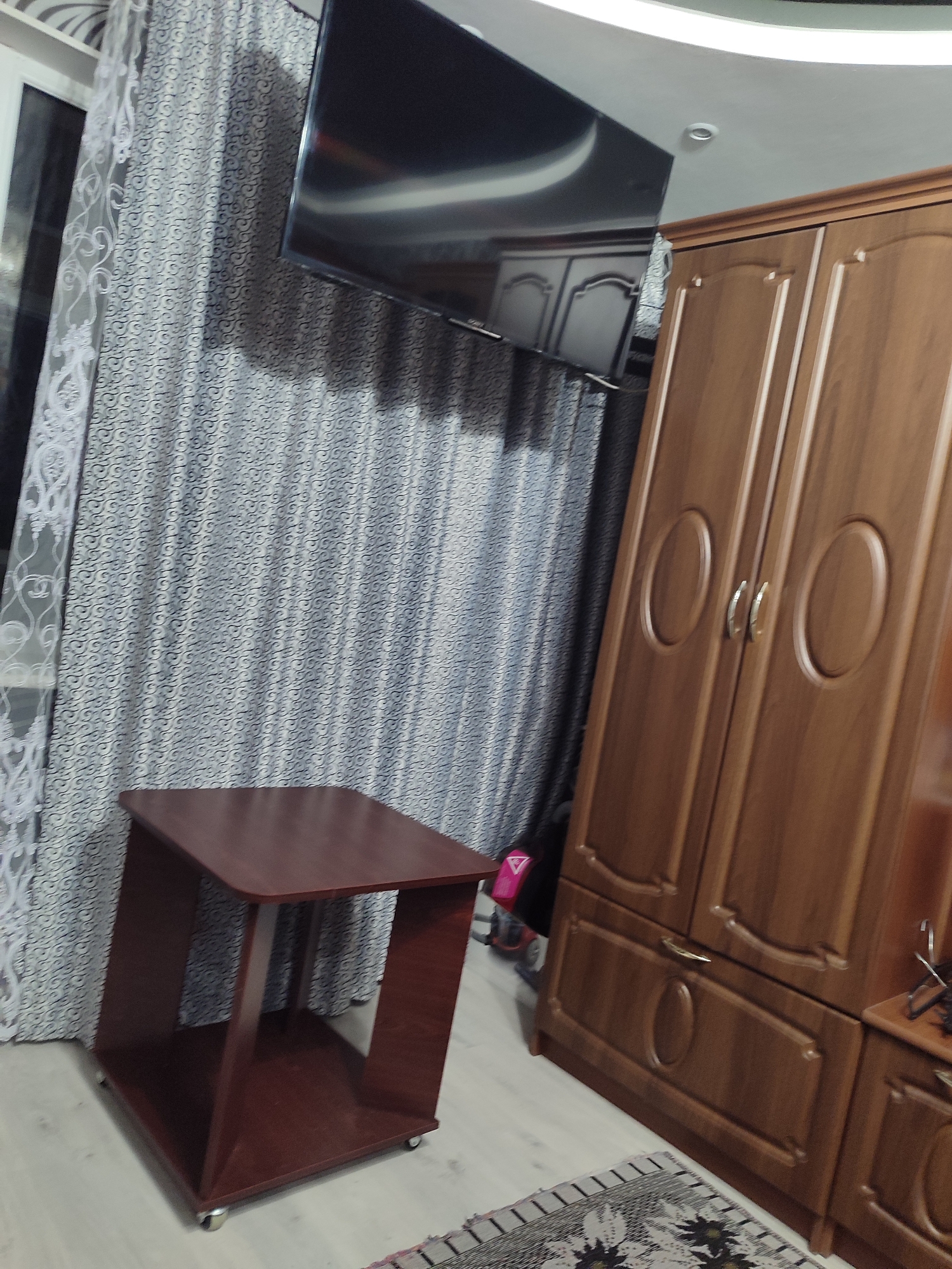 The image shows a room interior. The room contains a large wooden wardrobe with two doors, a small, dark brown side table on wheels, and a television mounted on the wall. The wardrobe has an ornate design with oval-shaped indentations on the doors and decorative trim. The side table is a simple, modern design with an open shelf. The television is a flat-screen and appears to be quite large. The room is decorated with patterned curtains. The flooring appears to be laminate or hardwood. There is also a rug visible on the floor. The lighting in the room is a mix of natural light from the window and artificial light from the ceiling. The overall impression of the room is one of a bedroom or living area. There is no indication of the location being in Mykolaiv, Ukraine.
