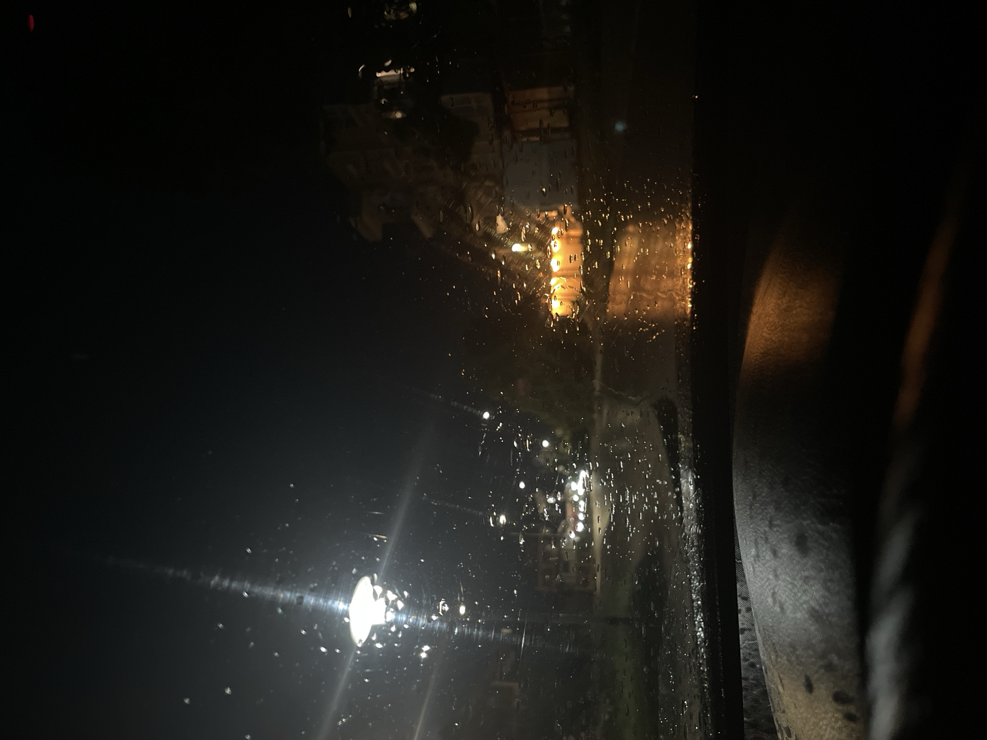 The image shows a view through a rain-streaked window at night. Out of the window, there are streetlights and faint building lights, likely indicating a street scene. The location is specified as Byala, Bulgaria. There are no people visible. The environment is dark and wet. The dominant objects are the window, raindrops, streetlights, and buildings. No actions or interactions are visible.
