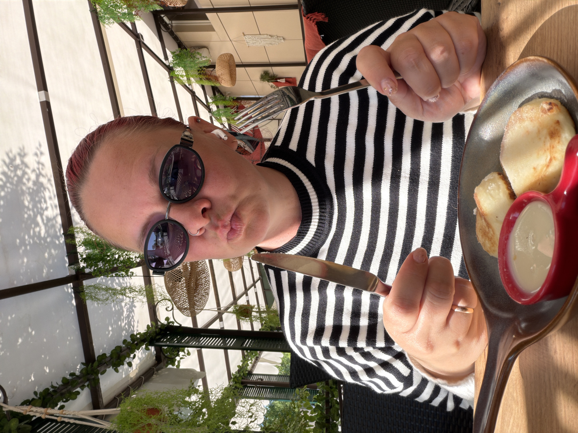 A person is photographed in what appears to be an outdoor dining area. The individual is holding a fork and knife, seemingly ready to eat a meal that includes small, round pancake-like items served on a rustic plate with a red ramekin of white sauce. The person is wearing sunglasses and a black and white striped sweater. The outdoor setting suggests a cafe or restaurant with overhead coverings and hanging plants. While the location cannot be verified to be Chaikovsky, Russia, the style of the food and the atmosphere of the dining area are consistent with a European or Russian setting.