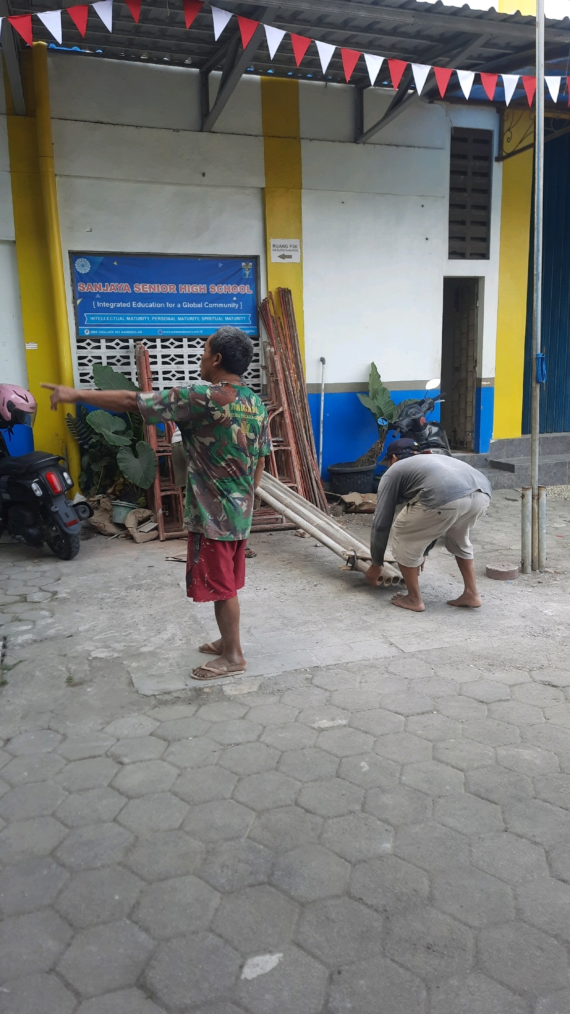 The image depicts an outdoor scene at Sanjaya Senior High School in Nanggulan, Indonesia. Two men are present. One man, wearing a camouflage t-shirt and red shorts, is standing and pointing to the left. The second man, in a grey long-sleeved shirt and light-colored shorts, is bent over and appears to be handling a pile of metal pipes. The environment includes a building with yellow and white walls, decorated with red and white triangular flags along the top. A sign for "SANJAYA SENIOR HIGH SCHOOL" and "RUANG PIK" is visible on the building. Construction materials, including scaffolding poles, are scattered on the ground. A motorcycle is parked to the left of the frame. The ground is paved with hexagonal concrete blocks.