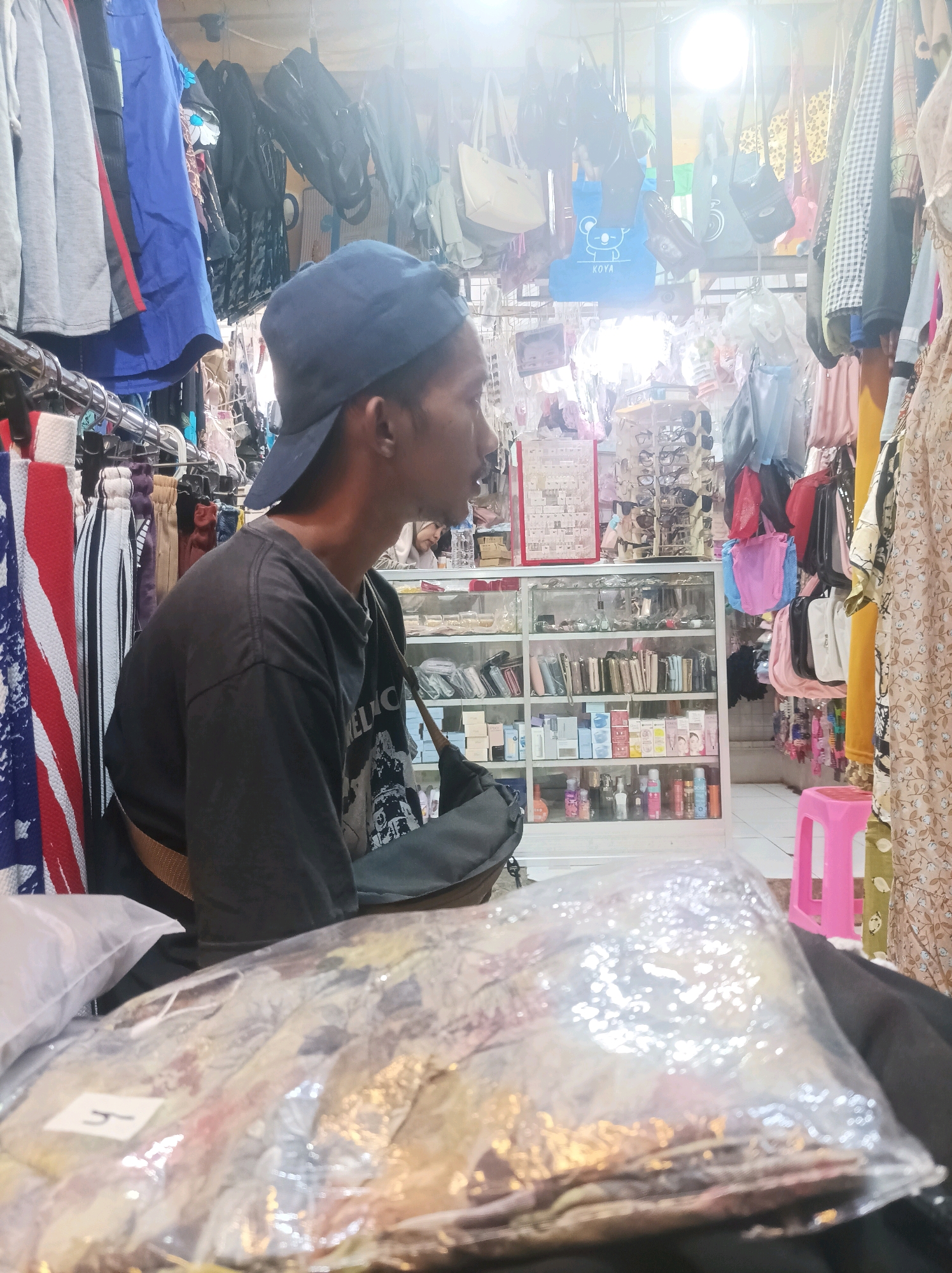 In a retail stall in Sumuragung, Indonesia, a young man wearing a dark blue baseball cap and a black t-shirt is seated. He is looking to his right, away from the camera. The stall is filled with merchandise, including clothing, bags, wallets, and cosmetics displayed on shelves and racks. In the foreground, a plastic-wrapped bundle of fabric with a visible label marked with the letter "H" rests on a surface. The lighting is bright, suggesting an indoor, possibly market, environment.