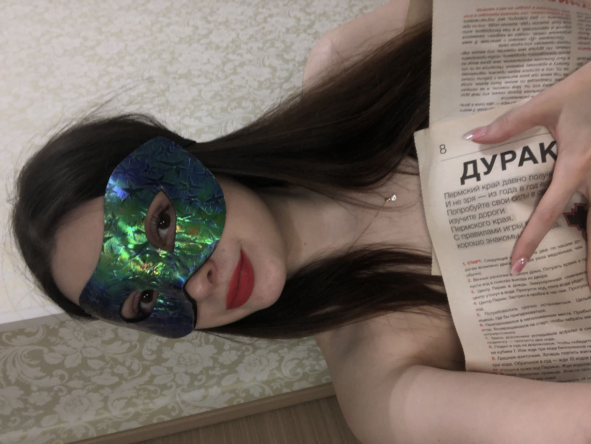 The image shows a person, likely a woman, lying down indoors in what appears to be Noviy, Russia. She is wearing a shimmering iridescent blue and green mask that covers the upper part of her face, revealing one eye looking directly at the camera. Her lips are painted with bright red lipstick, and her long, dark hair is spread around her. A delicate necklace with a small, sparkling pendant is visible on her chest.
She is holding an open book or magazine with her right hand. Her nails are manicured with a light, glossy finish. The visible text on the page is in Russian. A prominent heading reads "ДУРАК" (Fool) above a smaller segment of text that mentions "Пермский край" (Perm Krai), "дороги" (roads), and "правилами игры" (game rules), implying a localized game or guide. A numbered list outlines various scenarios or steps, such as "СТАРТ" (Start), "Вечные раскопки" (Eternal excavations), "Центр Перми в дождь" (Perm center in rain), "Застрял в пробке" (Stuck in traffic jam), "Припарковался в неположенном месте" (Parked illegally), "Подал в суд на дорожников" (Sued road workers), and "Гаишник-взяточник" (Traffic cop-bribe taker). The page number "8" is clearly visible. The background features a light-colored, patterned wallpaper and a wooden floor border. The image has a direct, engaging feel, likely a posed photograph.