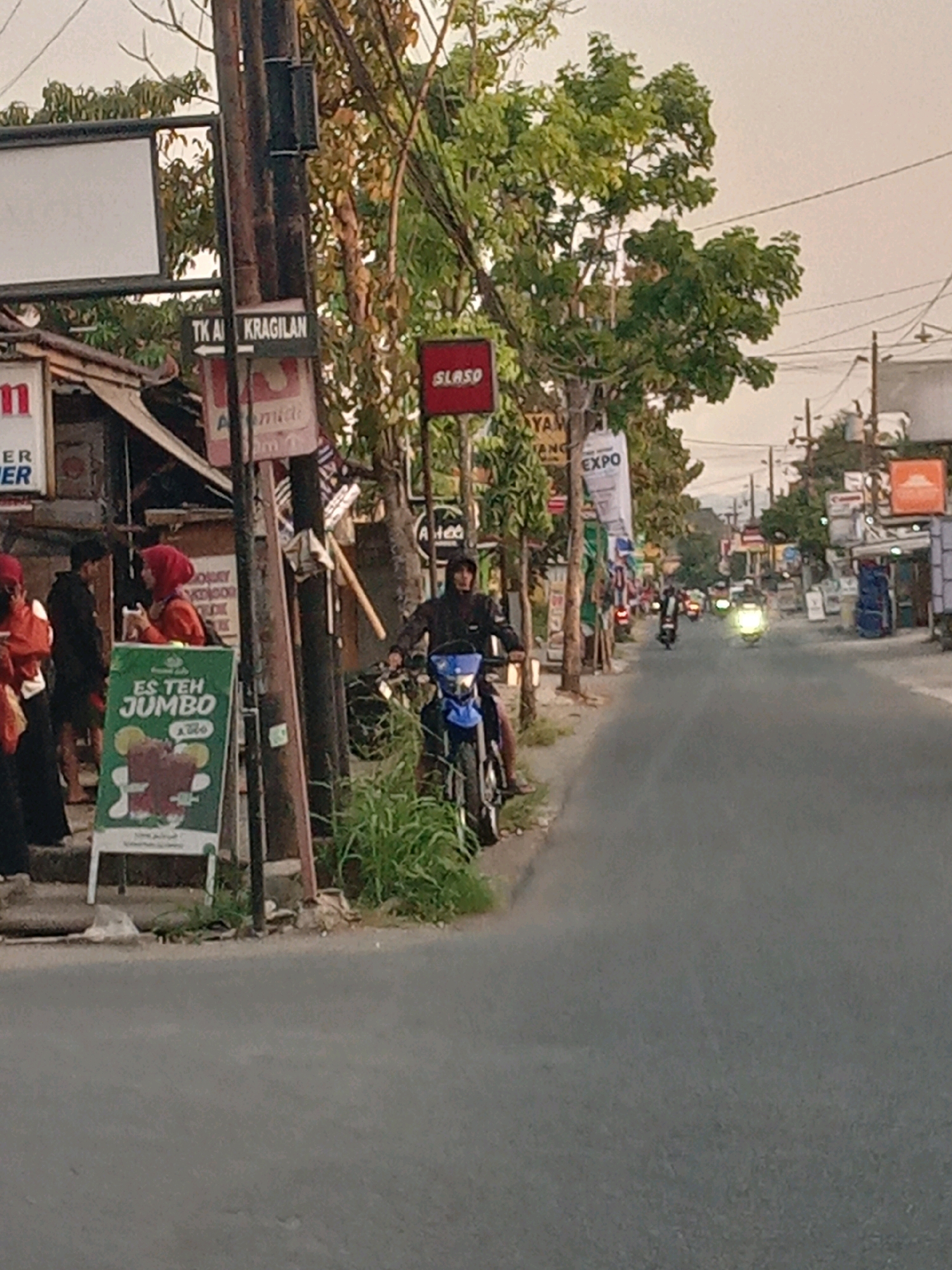 The image depicts a street scene in Banguntapan, Indonesia, likely during the daytime given the lighting conditions. In the foreground, a person on a blue motorcycle is visible, wearing a dark hooded jacket. The motorcycle has a prominent headlight. To the left of the road, a green A-frame sign advertises "ES TEH JUMBO" (Jumbo Iced Tea) with an illustration of a large cup of iced tea. Several individuals, some wearing headscarves, are present near a building on the left side of the frame, suggesting commercial activity.
A wooden utility pole stands prominently, displaying various signs, including one that reads "TK A KRAGILAN" (Kindergarten Kragilan) with an arrow pointing left, and another red sign with the word "SLASO". Further down the street, other motorcycles and scooters are in motion, indicating a moderately busy road. Buildings line both sides of the street, some appearing to be shops. The sky is overcast, with a pale, diffused light. The overall environment suggests a typical Indonesian urban or semi-urban street.