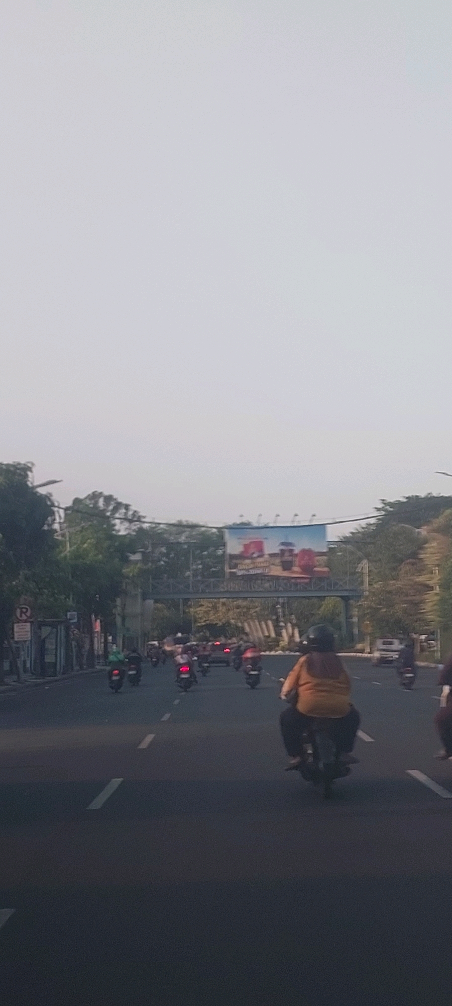 The image captures a street scene in Surabaya, Indonesia, likely during the daytime under an overcast sky. A busy road is filled with a multitude of motorcycles, suggesting typical traffic conditions in the city. In the foreground, a person on a motorcycle is visible, wearing a yellow top and dark pants, facing away from the camera. Further down the road, many other motorcycles are seen, some with riders and some without clear occupants due to motion blur. There's a pedestrian bridge spanning across the road in the mid-ground, and a large billboard is mounted on it, displaying advertisements for beverages. Trees line the sides of the road, and various buildings and structures are present, including what appears to be a bus stop or information kiosk on the left with a red circular sign indicating a restriction. The overall impression is one of an active urban environment with significant motorcycle usage.