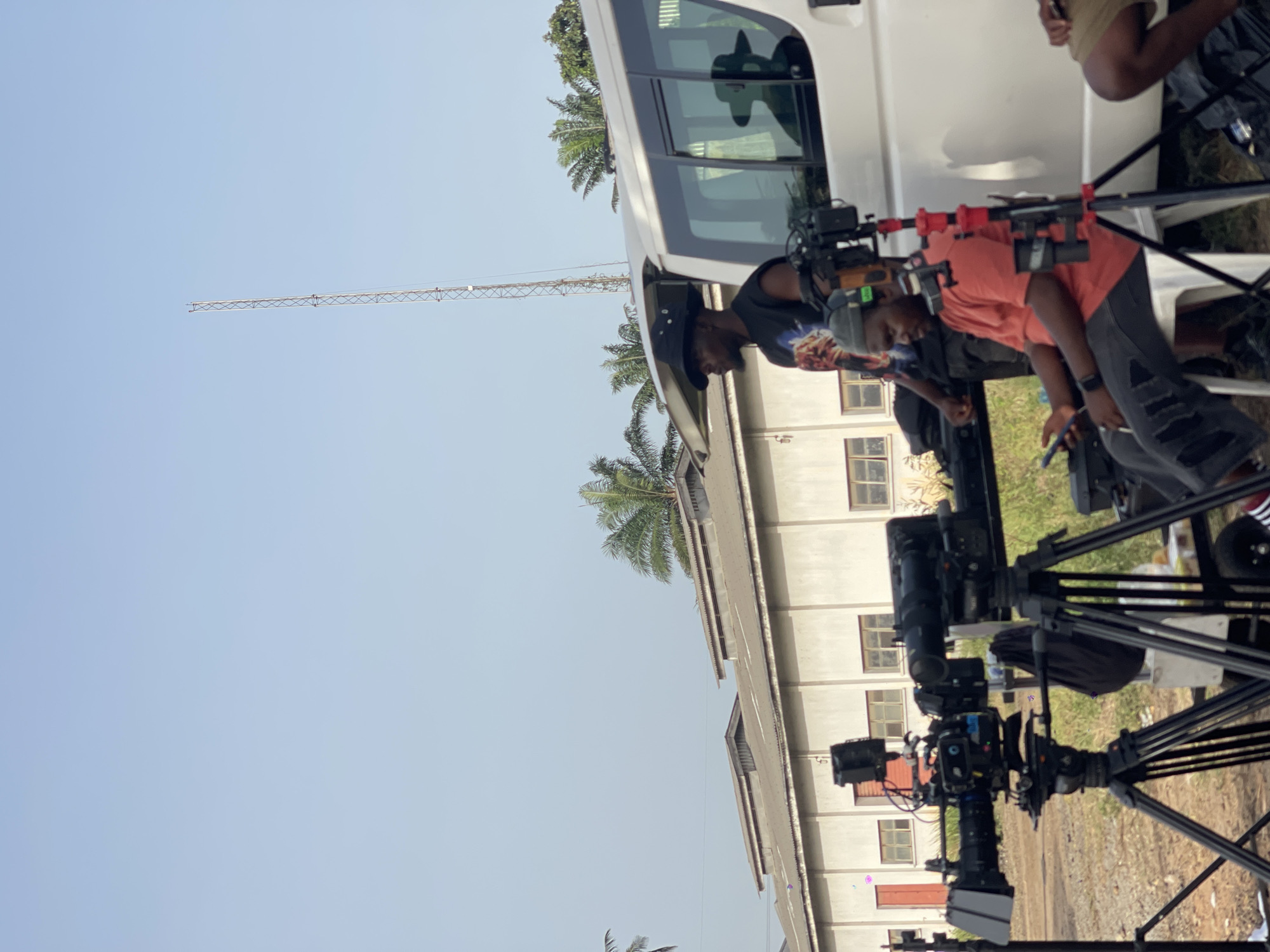 A medium, outdoor shot captures a film set in an unidentified urban location. A white van is partially visible on the left side of the frame, with two dark-skinned individuals leaning into the vehicle's open side door. The person on the left wears a black shirt and hat, while the other is dressed in a peach-colored shirt. Two professional camera setups on tripods stand in the foreground. In the background, a single-story building with multiple windows is visible. Above, a clear sky contrasts with the surrounding structures, and a tall metal communication tower is seen in the distance. Palm trees add a touch of nature to the urban setting.