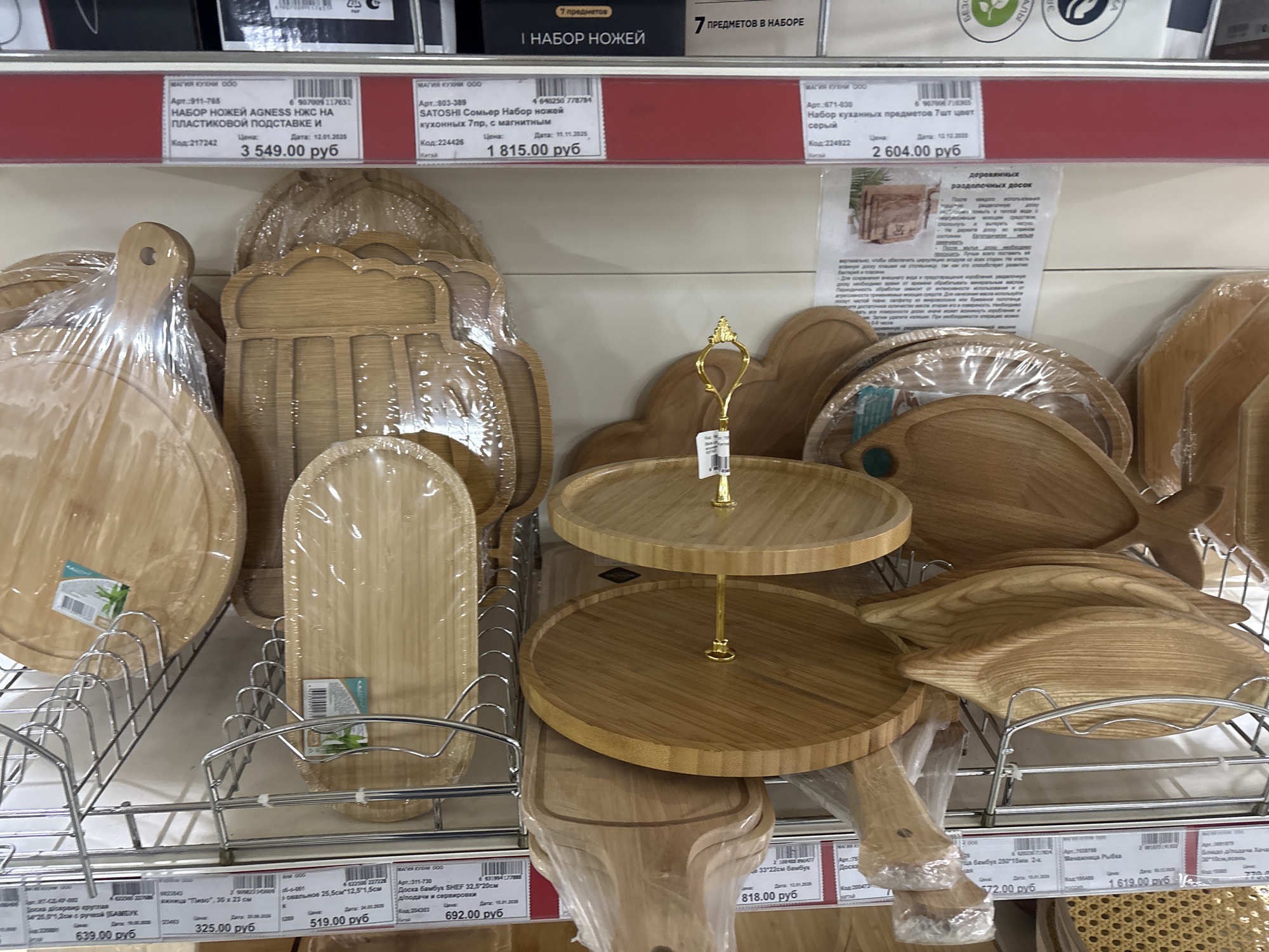The image shows a shelf in a store, likely in Krasnoyarsk, Russia, displaying various wooden kitchen items. These include cutting boards of different shapes and sizes, some wrapped in plastic. A two-tiered serving stand made of wood is also present. Price tags are visible below the items. There are no people present in the frame.