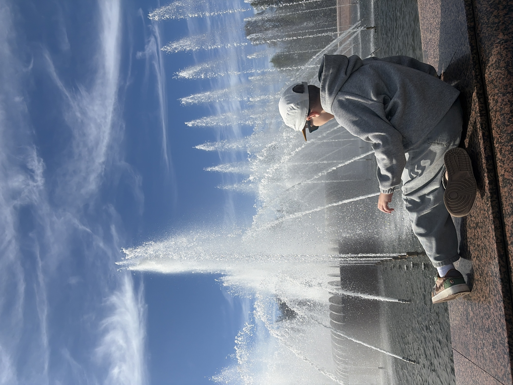 A young person leans over the edge of a large outdoor fountain, engrossed in watching the water jets under a bright blue sky. The image, presented in a portrait orientation but depicting a landscape scene, captures the individual from behind and to the side, highlighting their focused posture. They are dressed casually in a grey hooded sweatshirt, light grey trousers, a white baseball cap, and sneakers, with sunglasses resting on their cap.
The fountain features numerous powerful jets of water spraying upwards and outwards, creating a lively display of splashing water. These water plumes are set against a clear sky adorned with wispy white clouds. The person is positioned next to a dark, polished stone or granite ledge that forms the border of the fountain, with the water surface visible below.
The scene suggests a sunny day, likely midday or early afternoon, given the intensity of the light reflecting off the water. The overall mood is one of quiet observation and childlike curiosity. No discernible text is visible within the image. This tranquil moment takes place in Saint Petersburg, Russia.