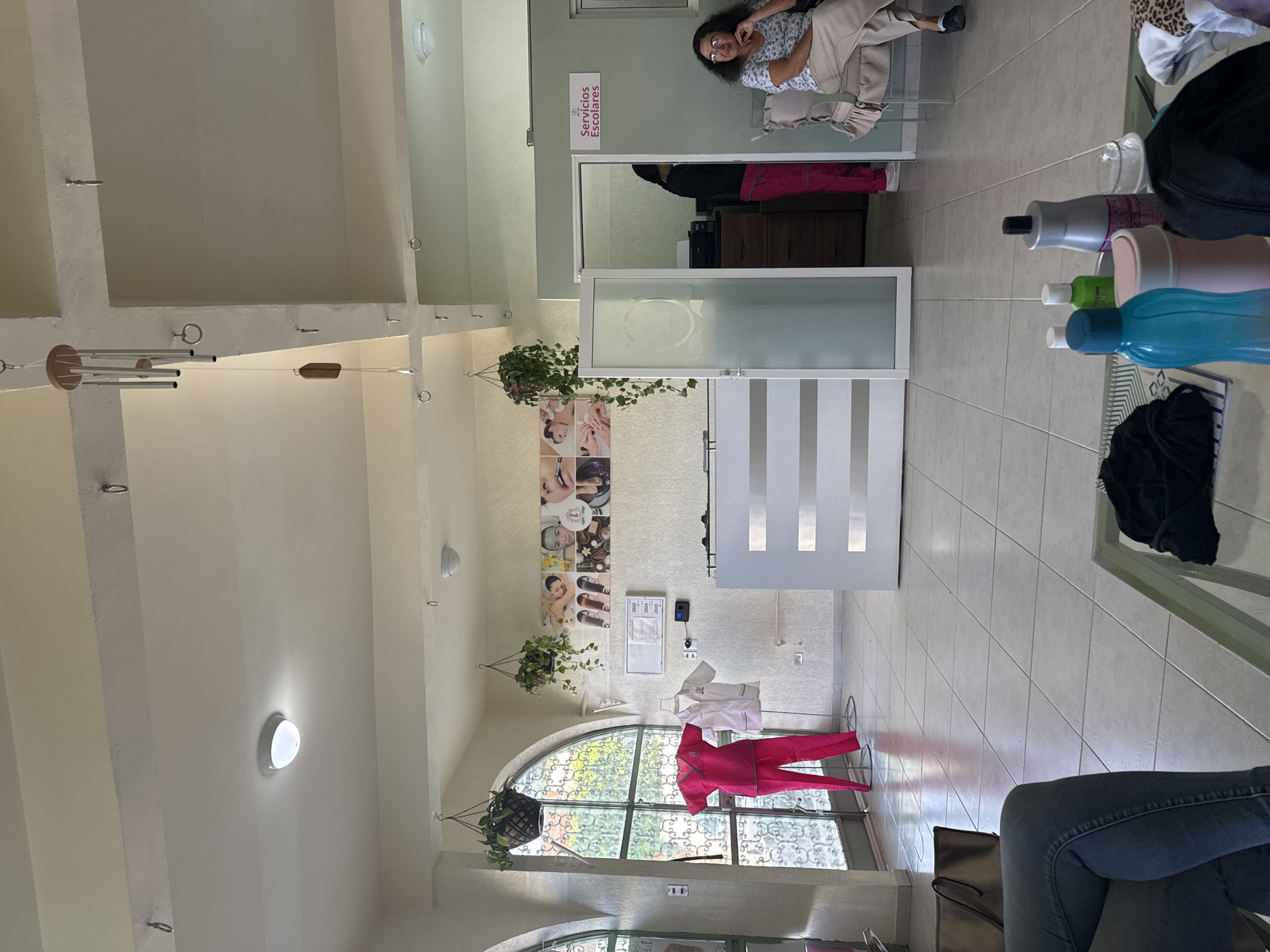 An indoor scene, possibly a vocational school or clinic in Puebla, Mexico, is captured from a low vantage point, showing various elements of the space. The tiled floor and light-colored walls suggest a clean and organized environment.
In the midground, a woman with dark hair and glasses is seated, looking towards the camera with a slight smile. She is wearing a patterned top and light-colored pants. Behind her, through a frosted glass door, a reflection shows another person in a black top and pink pants. To the right of the visible woman, a shelf or table holds several bottles, including shampoos, conditioners, and plastic water bottles.
The ceiling features square recesses and multiple circular lights. Hanging from the ceiling are wind chimes and two planters with green foliage. On the wall, several posters display images related to beauty treatments such as facials and manicures. Below these posters, a pink scrub-like uniform is displayed on a mannequin, and a white lab coat is shown on another. An arched window with decorative metalwork provides a view of outdoor greenery, indicating it is daytime.
A white sign prominently displays the text "Servicios Escolares" (School Services), suggesting an educational institution. Below it, a paper with "2023", "2024", and "2025" is visible. The legs of the person taking the photo are visible in the foreground, indicating they are seated. The overall atmosphere appears calm and functional.