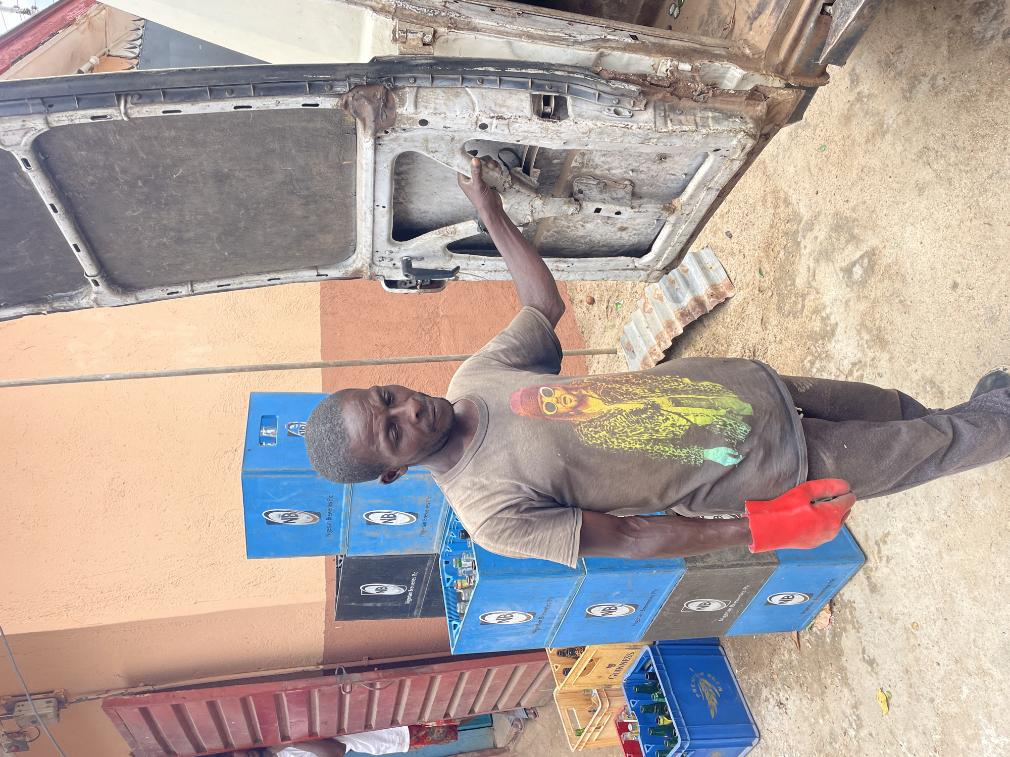 A man wearing a brown t-shirt with a yellow print stands to the left of a stack of blue and black crates branded with "Nigerian Breweries Plc," with some crates containing glass bottles, in what appears to be Jos, Nigeria. He is using his right hand to work on the interior of a car door, which is detached and leaning against the vehicle. He is wearing a red protective glove on his left hand. A piece of corrugated metal lies on the ground near the door. A tan-colored wall is visible behind the stack of crates, while a weathered and damaged red metal container is situated in the lower left. The ground is concrete with scattered debris.