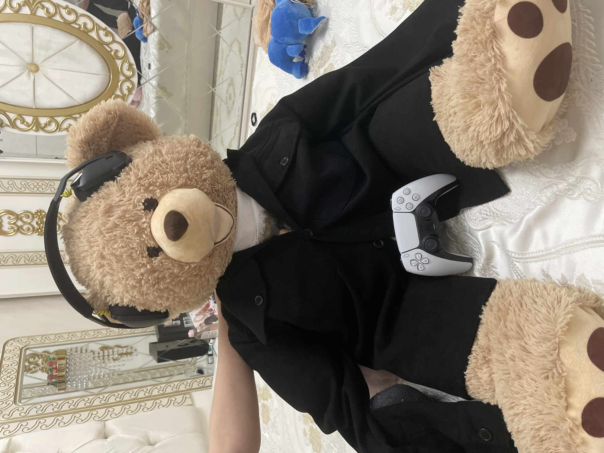 A large, light brown teddy bear, wearing black over-ear headphones and dressed in a black long-sleeved shirt and matching pants, is positioned on a white, textured bed or sofa. Its plush, light brown feet, adorned with dark brown spots, are visible towards the lower right. A white PlayStation 5 DualSense controller rests on the teddy bear's midsection. In the upper center, a small blue plush toy, resembling Stitch from Disney, lies on the white surface.
The setting is an indoor room, likely a bedroom, characterized by ornate white and gold furniture. A portion of a decorative headboard or furniture piece with gold trim is visible to the left. Below the teddy bear's head, a gold-framed mirror reflects what appears to be crystal or ornate light fixtures. The lighting suggests an indoor environment. The scene presents a whimsical, staged arrangement, implying the teddy bear is engaged in gaming. No visible text is present in the image. This scene is located in Ihtiman, Bulgaria.