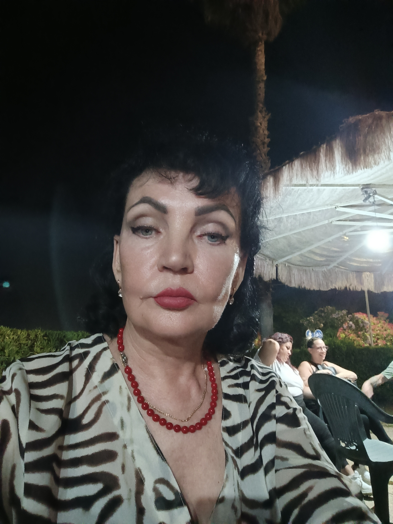 The image depicts a woman in the foreground, facing the camera, with her head and shoulders filling most of the frame. She has dark, styled hair, prominent makeup including red lipstick and eyeliner, and is wearing a white and brown animal-print garment. A red beaded necklace is visible around her neck. Behind her, in softer focus, are other individuals seated at tables under a large, white thatched umbrella structure. The setting appears to be an outdoor seating area at night, with some greenery visible. The overall atmosphere suggests a casual evening gathering. The image does not provide any specific details indicating it is in Giulianova, Italy.