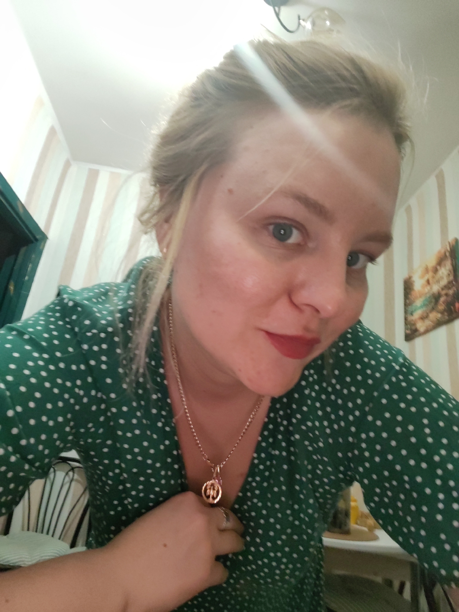 A woman with blond hair is in a room. She is wearing a green polka dot shirt and a gold necklace. Her left hand is placed on her chest. The background includes striped wallpaper, a green doorway, and a small painting. The image location cannot be determined to be Izhevsk, Russia, from visual information.