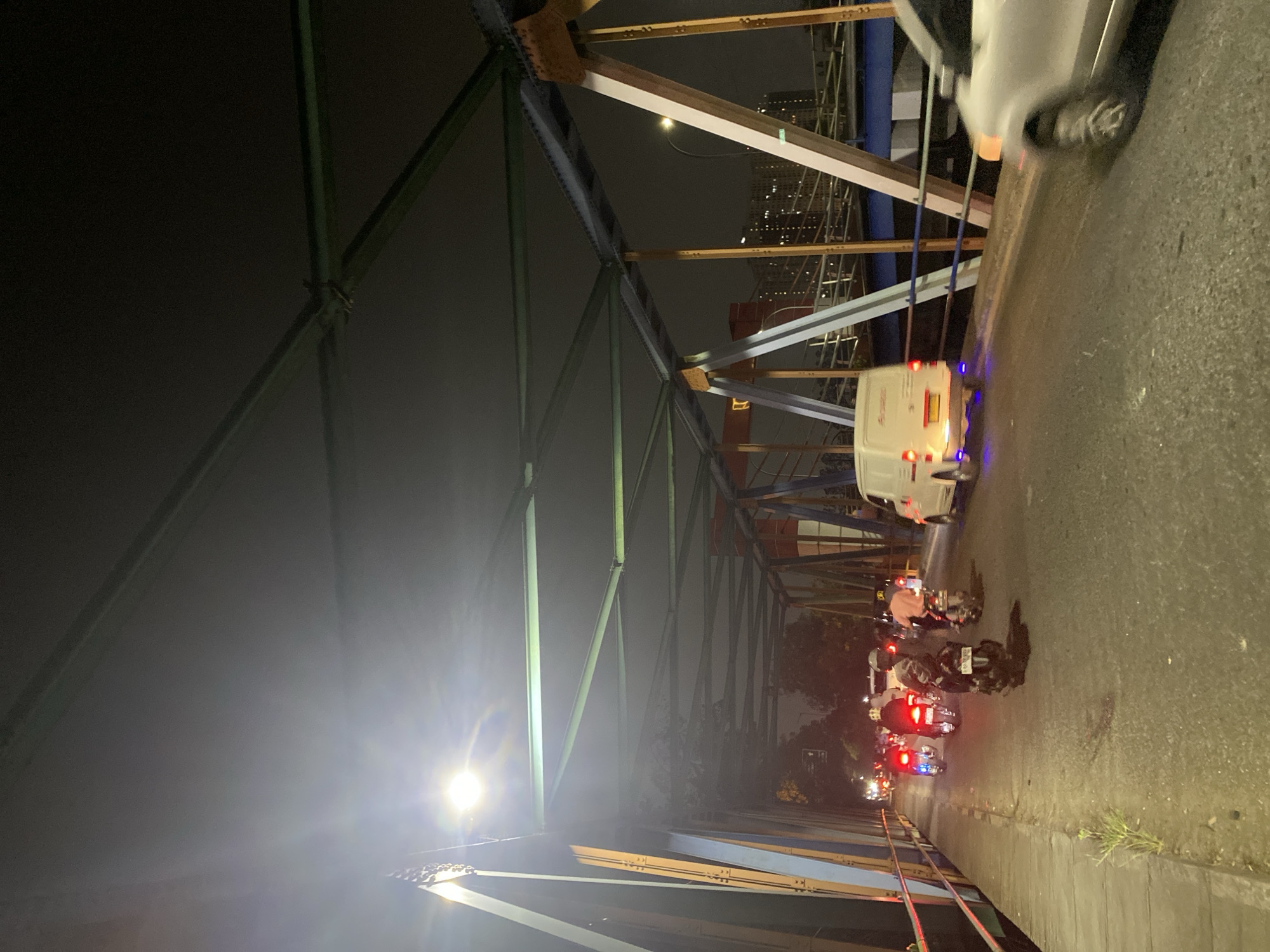 The image captures a nighttime view of a bridge in Tangerang, Indonesia, likely at an intersection or busy artery. The bridge features a prominent steel truss structure painted in green and accented with orange and blue structural elements. A bright street lamp illuminates the scene from the left side of the frame. The bridge deck is paved and shows signs of wear. Traffic is present, with several motorcycles in the foreground, their taillights glowing red, and one white van with its brake lights illuminated. A white car is captured in motion on the right side of the bridge, its front wheel blurred due to movement. In the background, a cityscape with illuminated buildings is visible, suggesting an urban environment. The overall atmosphere is dark, with artificial lighting from vehicles and street lamps creating stark contrasts.