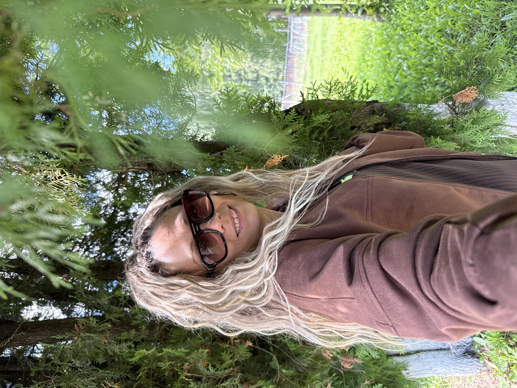 In Zhukovo, Russia, a woman with long, curly blonde hair and sunglasses stands in front of a cedar tree, partially obscured by its branches. She is wearing a brown jacket and a brown shirt. The background shows a lush green lawn and a fence. The lighting suggests it is daytime, and the overall scene is outdoors and natural.