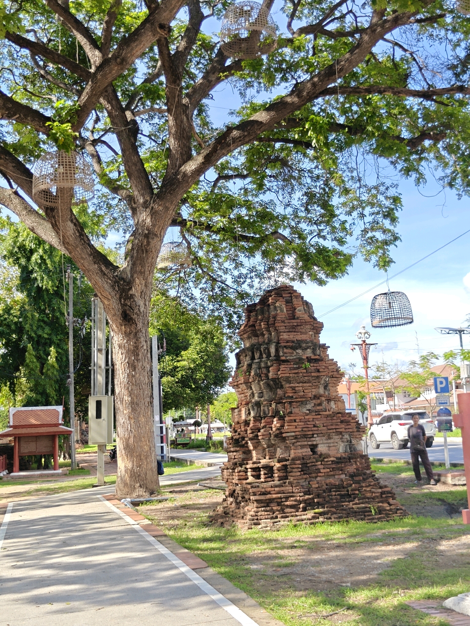 An outdoor, daytime, medium shot captures a brick ruin in Phra Nakhon Si Ayutthaya City Municipality, Thailand. The ruin, made of weathered, red bricks, is situated in front of a large tree with a textured trunk and green foliage. Hanging from the tree branches are two decorative woven baskets. A paved walkway with a white line borders the grassy area where the ruin and tree stand. In the background, a person wearing dark clothing walks on the far side of a street. Several cars are parked nearby, and buildings with terracotta roofs are visible in the distance. The sky is a clear blue with some wispy clouds.