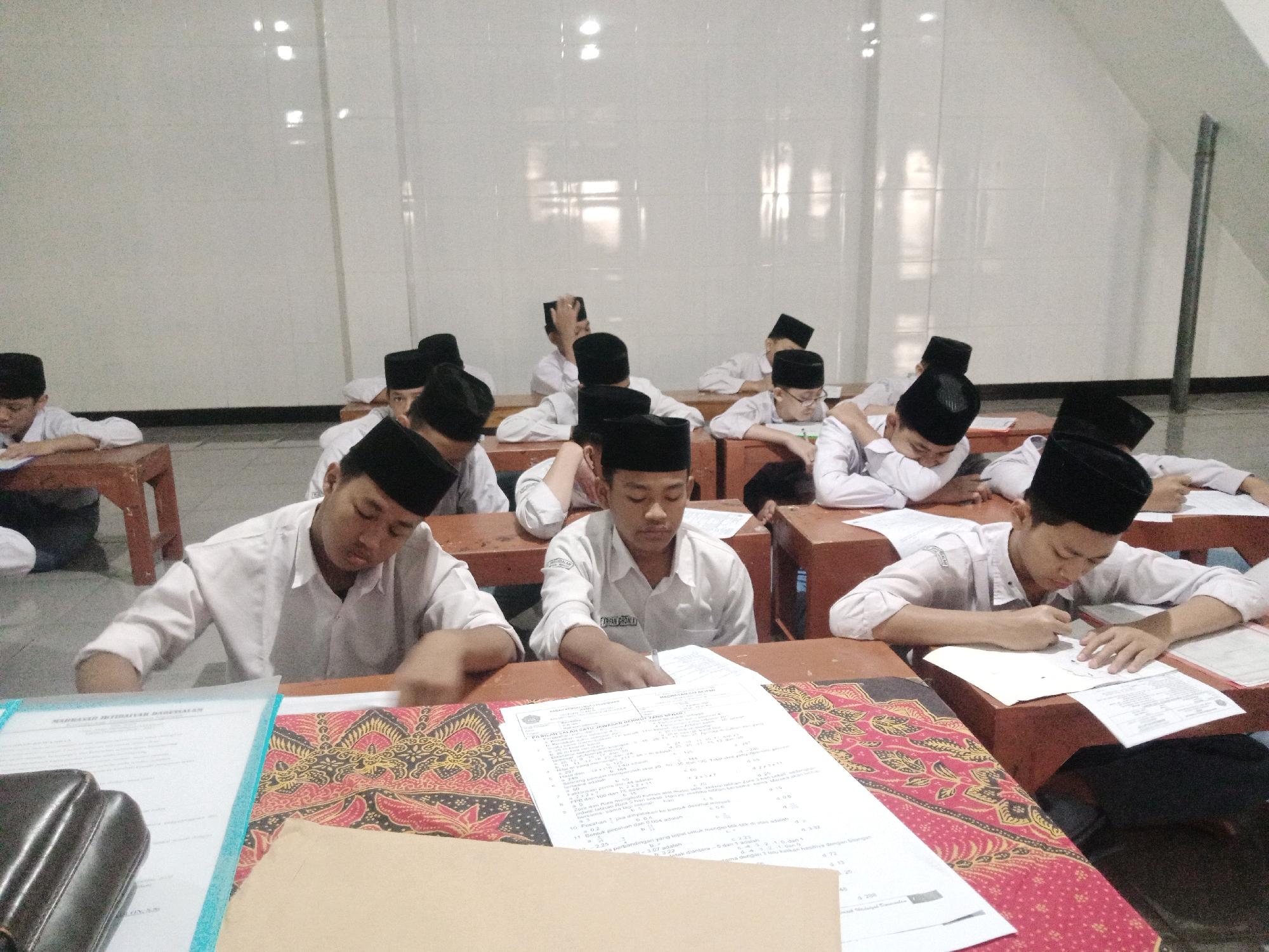 In a classroom with white tiled walls, students wearing white shirts and black skullcaps sit at wooden desks. Most of the students are looking down at papers on their desks, suggesting they are taking a test or completing assignments. One student in the foreground is actively writing on a paper. Another student in the middle of the frame appears to be looking up or thinking. The desks are arranged in rows, and the lighting in the room seems to be from overhead fluorescent lights. There are also decorative cloths with patterned designs on some of the desks. The overall scene depicts a typical classroom setting focused on academic work.